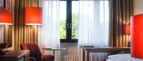 Habitación de hotel con cama, escritorio, silla, radiador y ventana con cortinas.