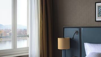 A hotel room with a large window, a bed with white linens, a blue headboard, and a lamp on a bedside table.
