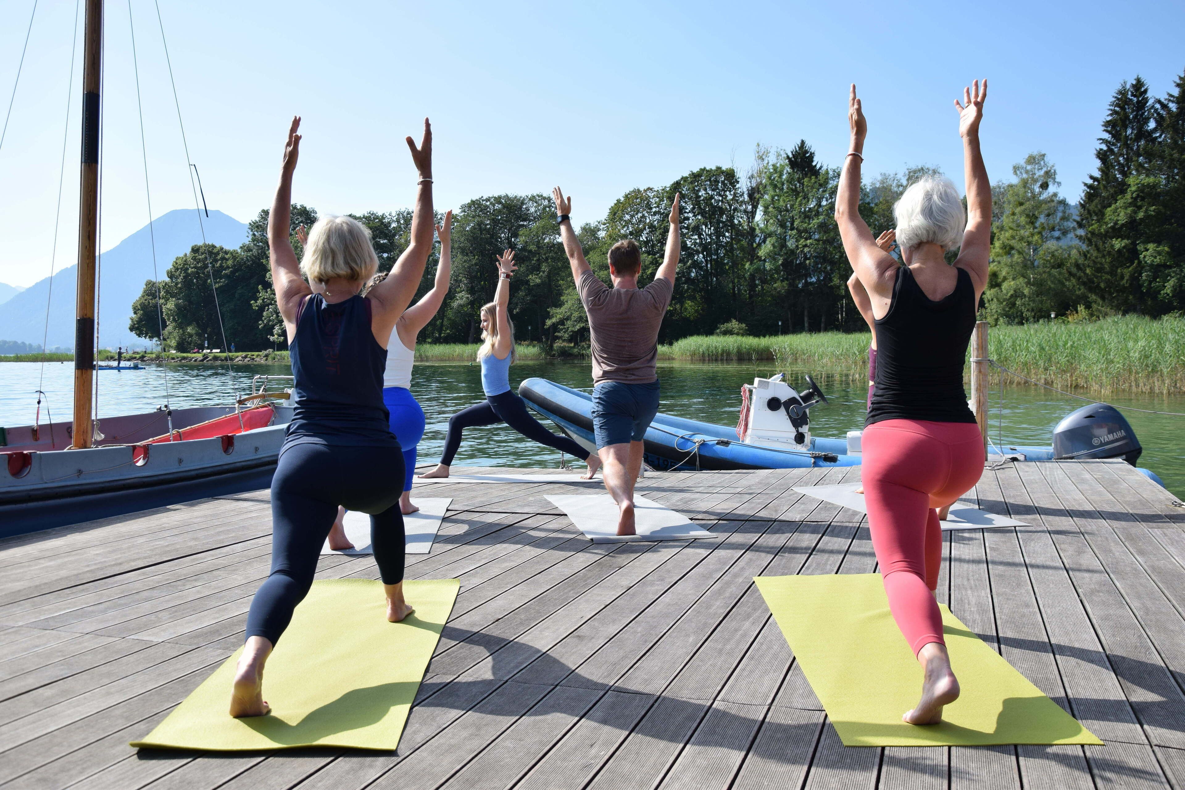 A group of people performing yoga on a wooden deck by a lake with boats in the background.
