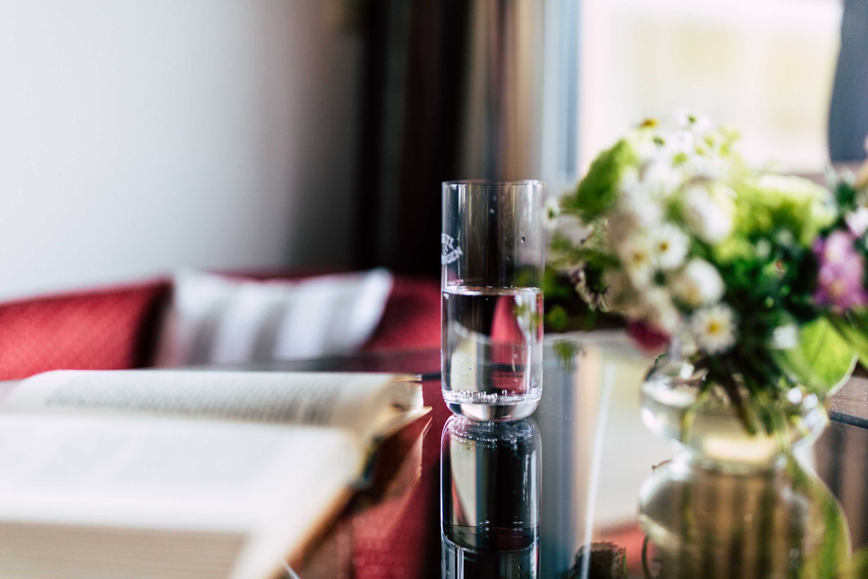 A clear glass of water on a table, a book, and a vase with flowers.