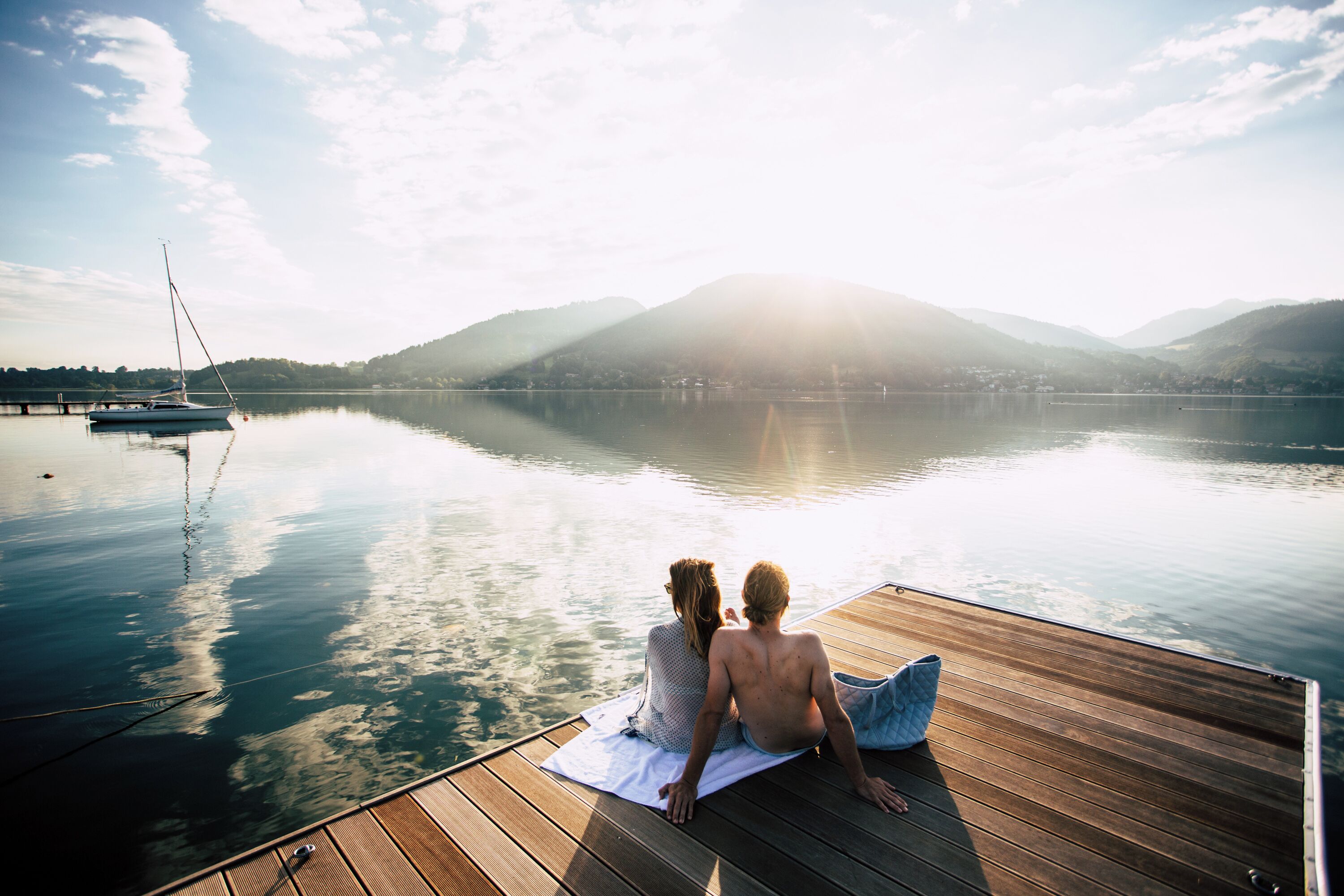 Two people sitting on a wooden dock with a view of a calm lake and mountains in the background.