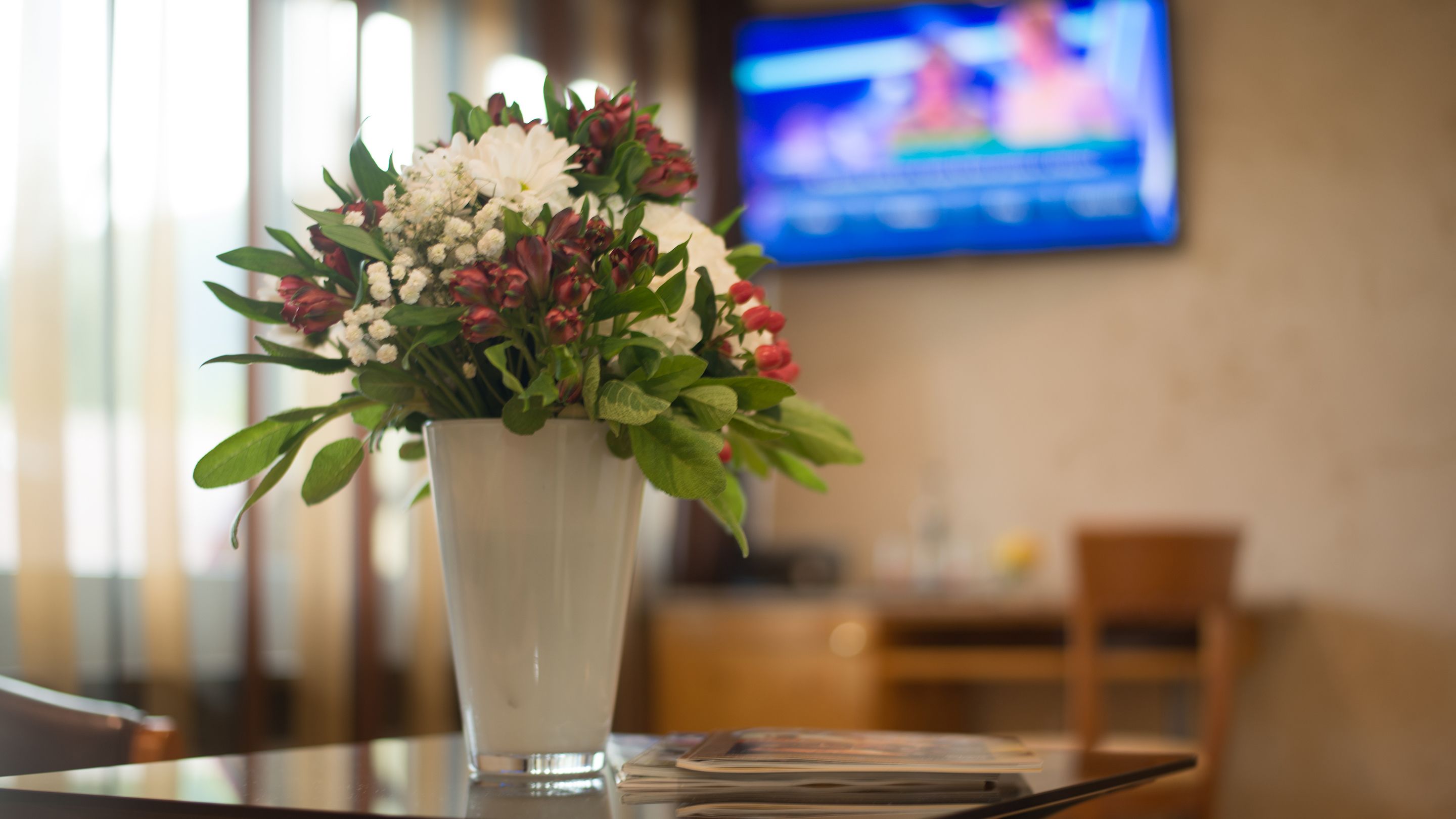 Un jarrón blanco con un arreglo floral sobre una mesa en una habitación, con un televisor al fondo.