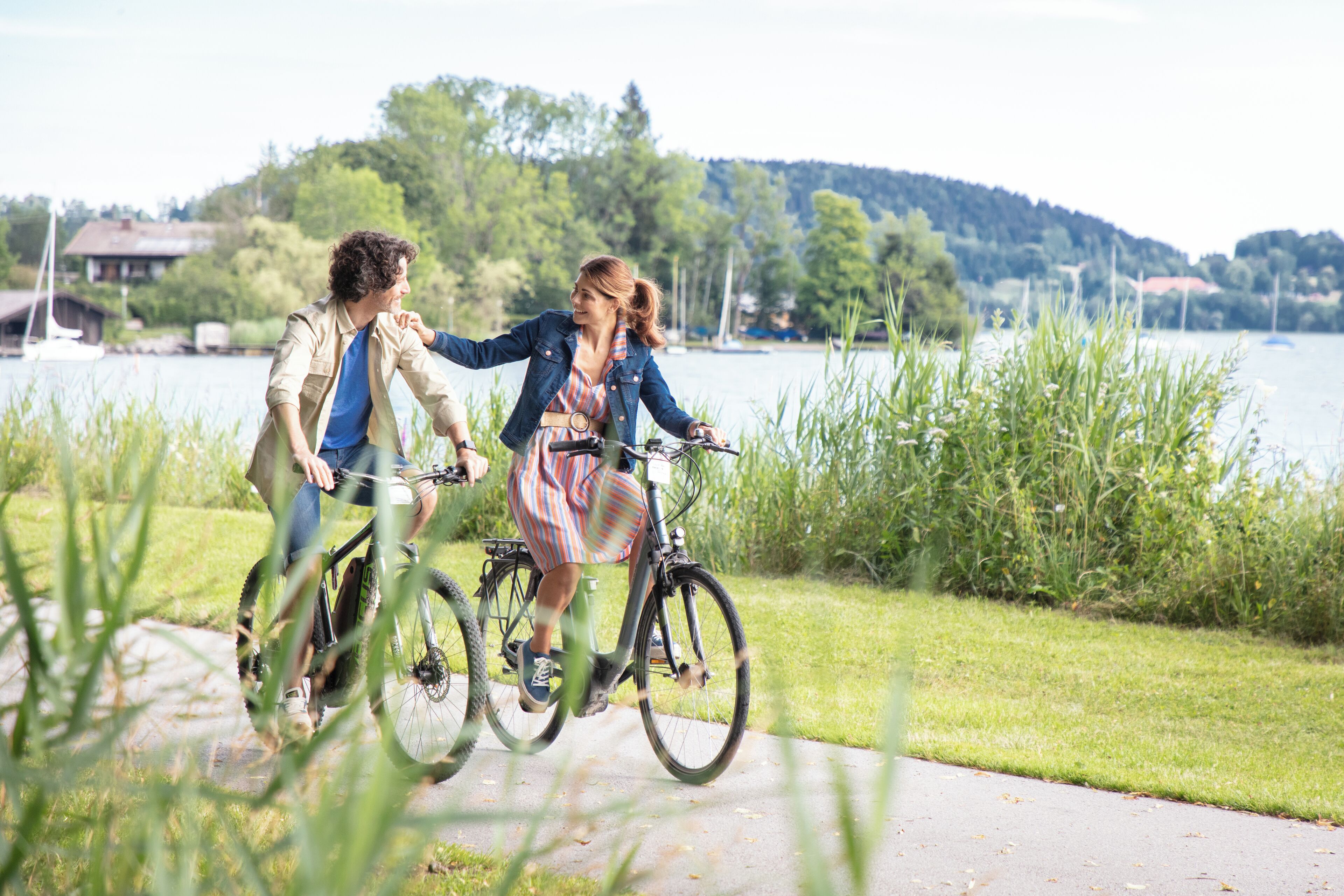 Two people riding bicycles on a paved path near a body of water with boats in the background.