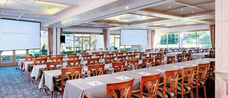 A conference room with rows of tables and chairs, large windows, and a projector screen displaying "Herzlich Willkommen".