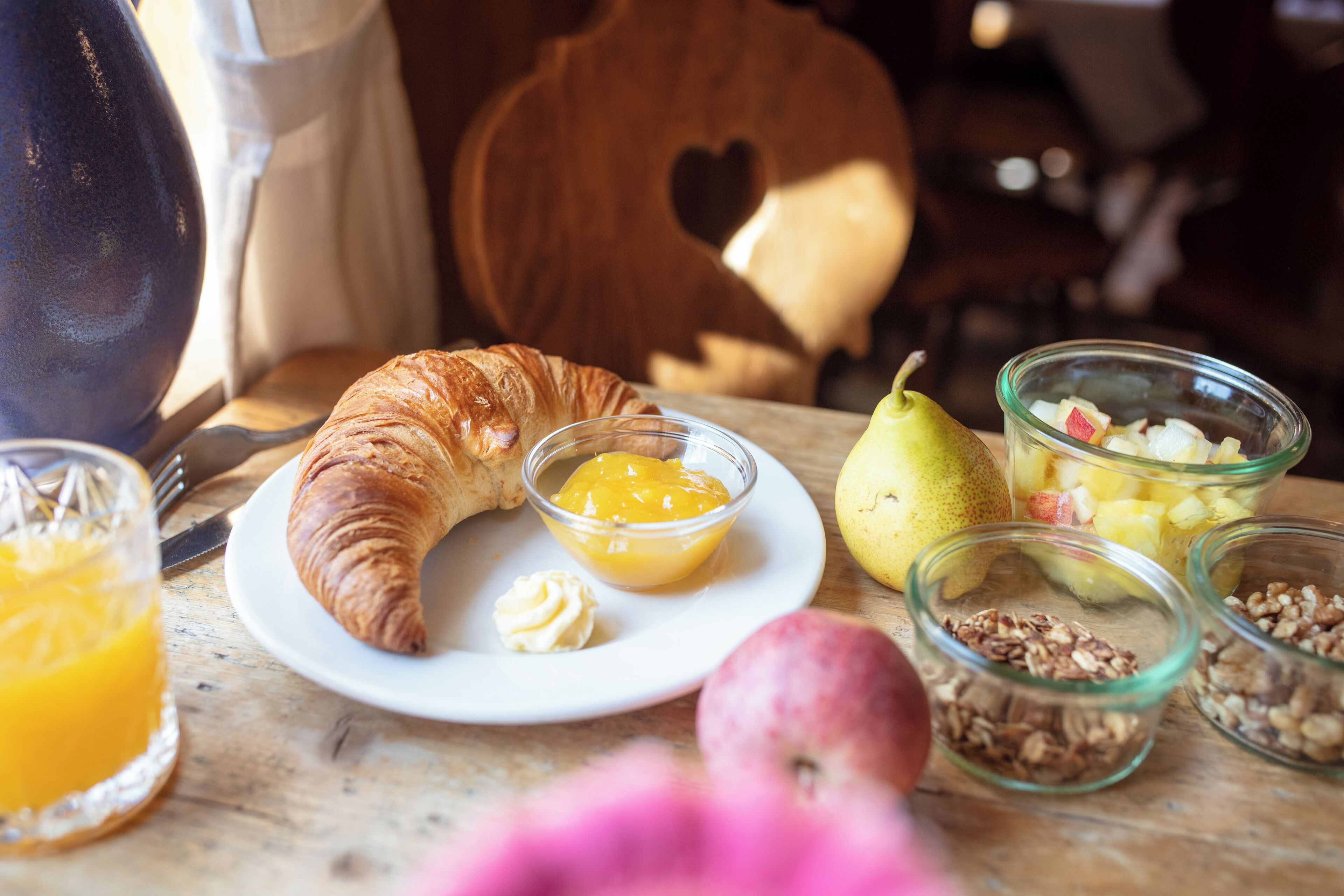 Un desayuno con croissant, zumo de naranja, pera y diferentes frutas y frutos secos sobre una mesa de madera.