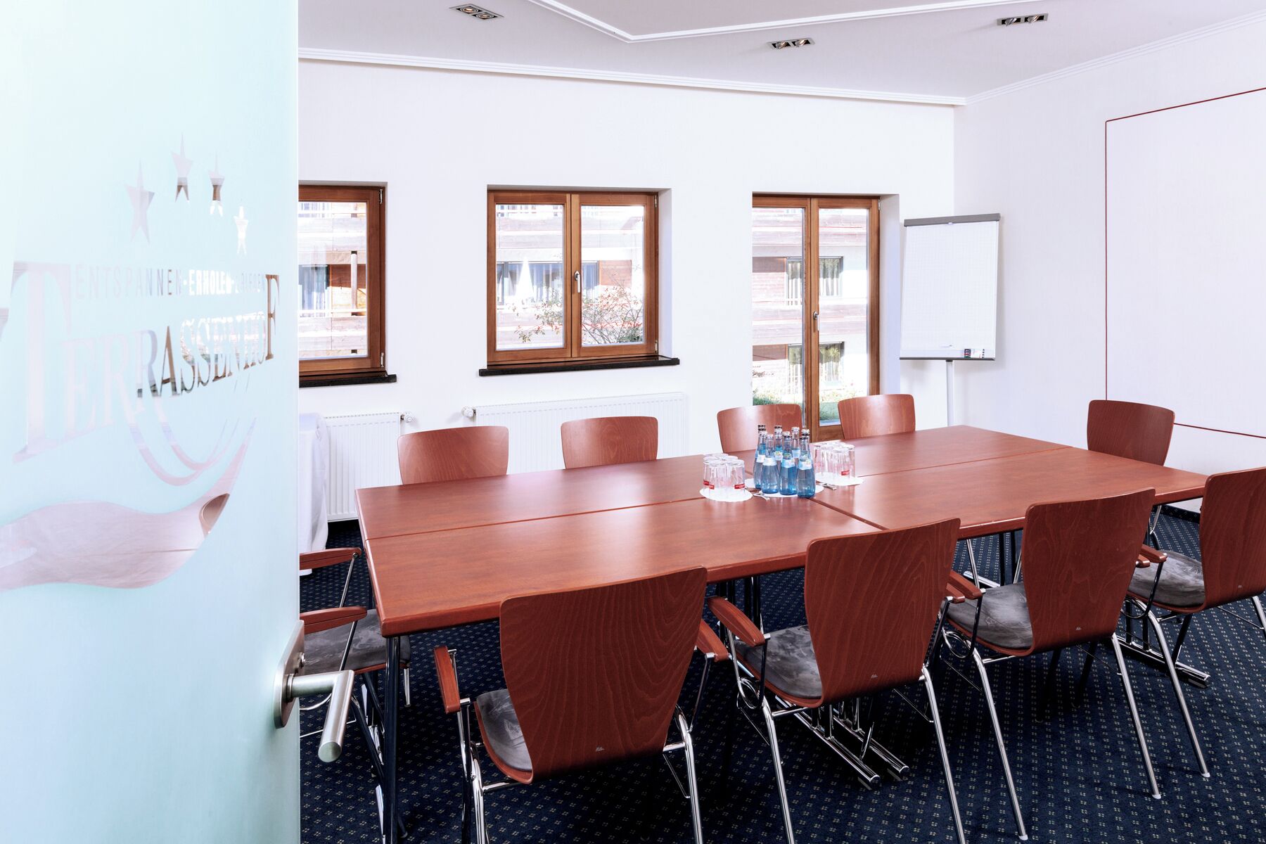 A conference room with a long table, six chairs, a whiteboard, and windows with a view of buildings outside.