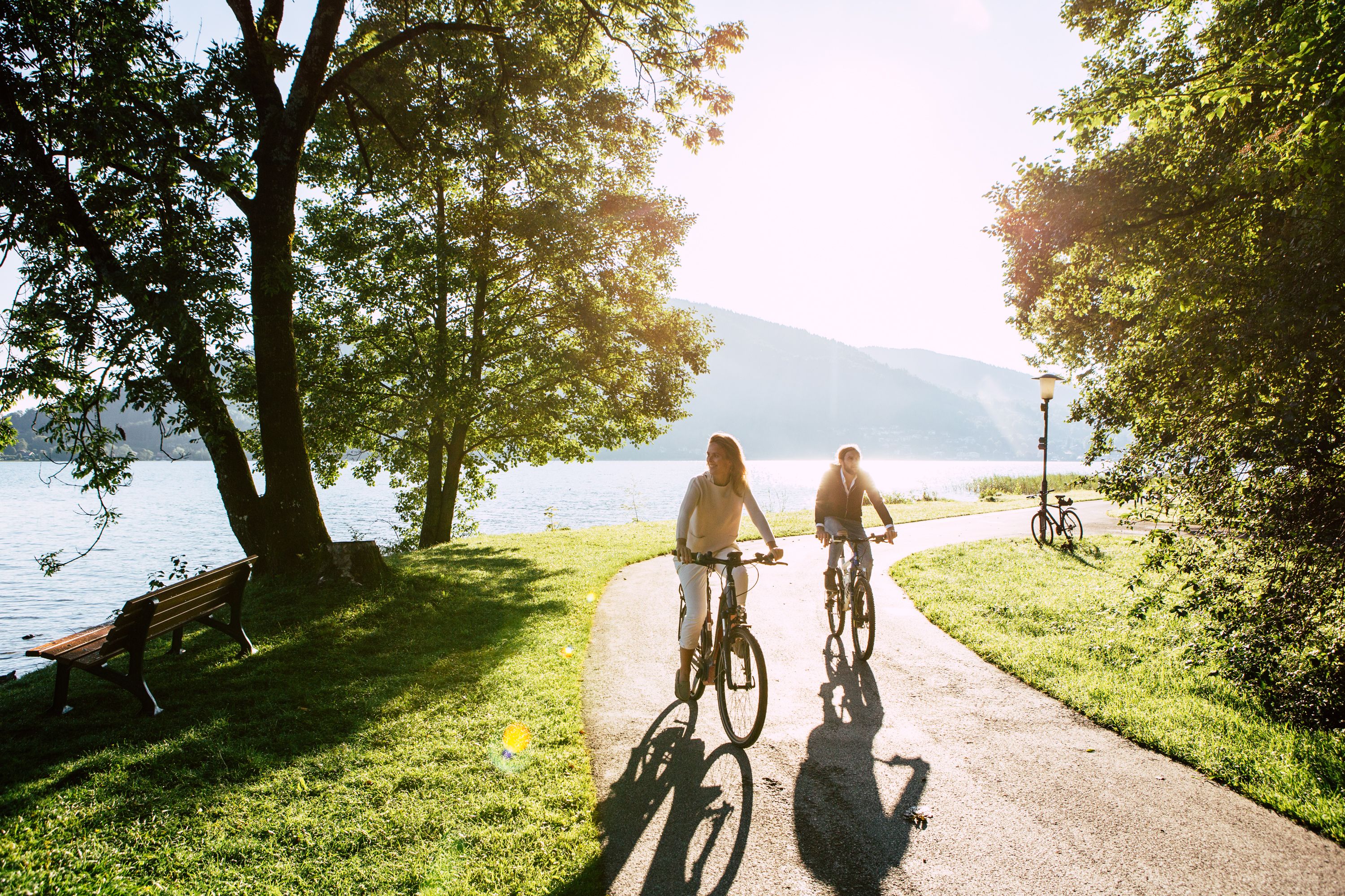 A paved path with benches and bicycles, surrounded by trees and a lake.