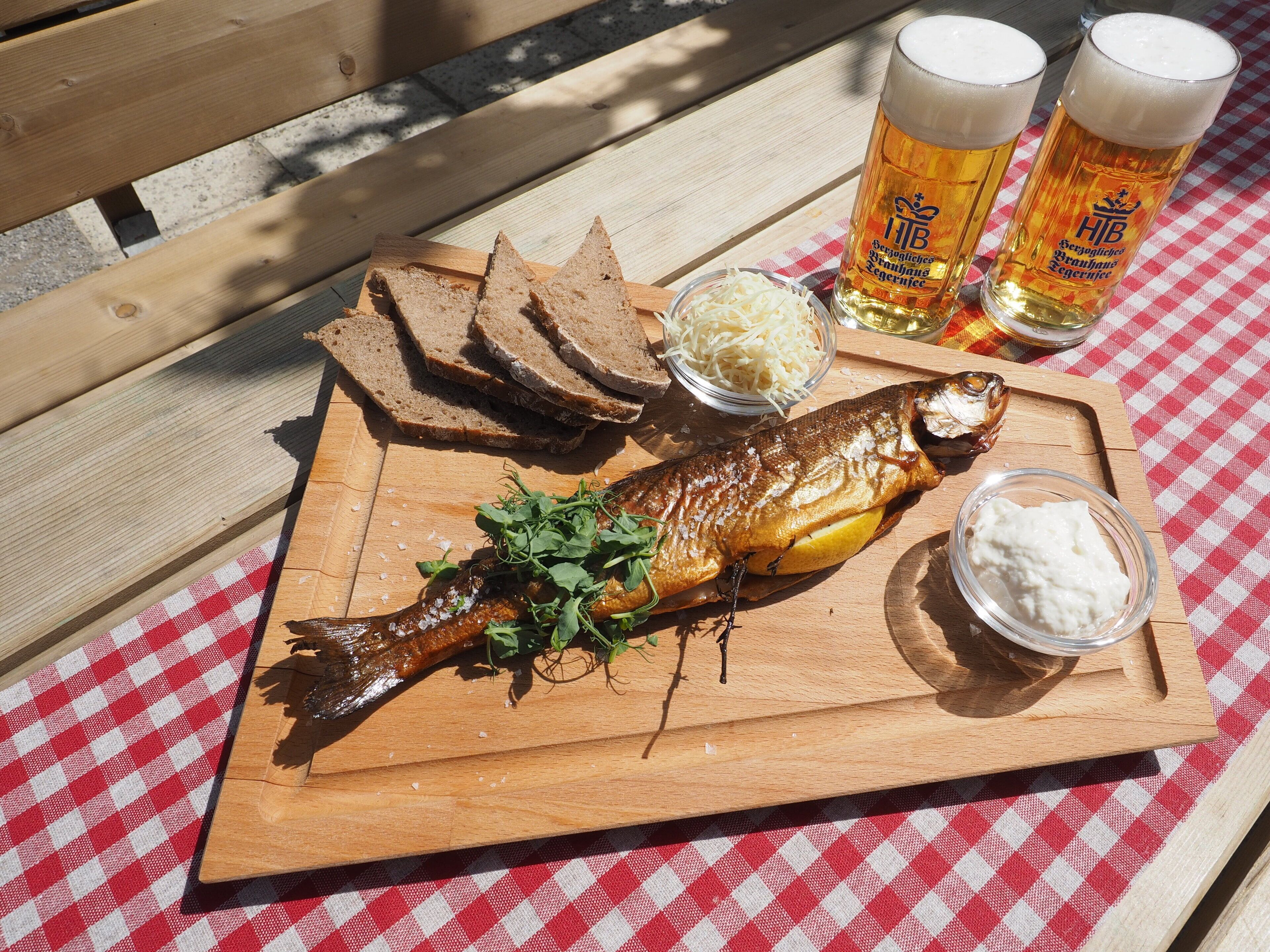 Una tabla de madera con un pescado asado, pan en rebanadas, queso rallado y un recipiente pequeño con salsa, todo sobre un mantel a cuadros rojo y blanco.