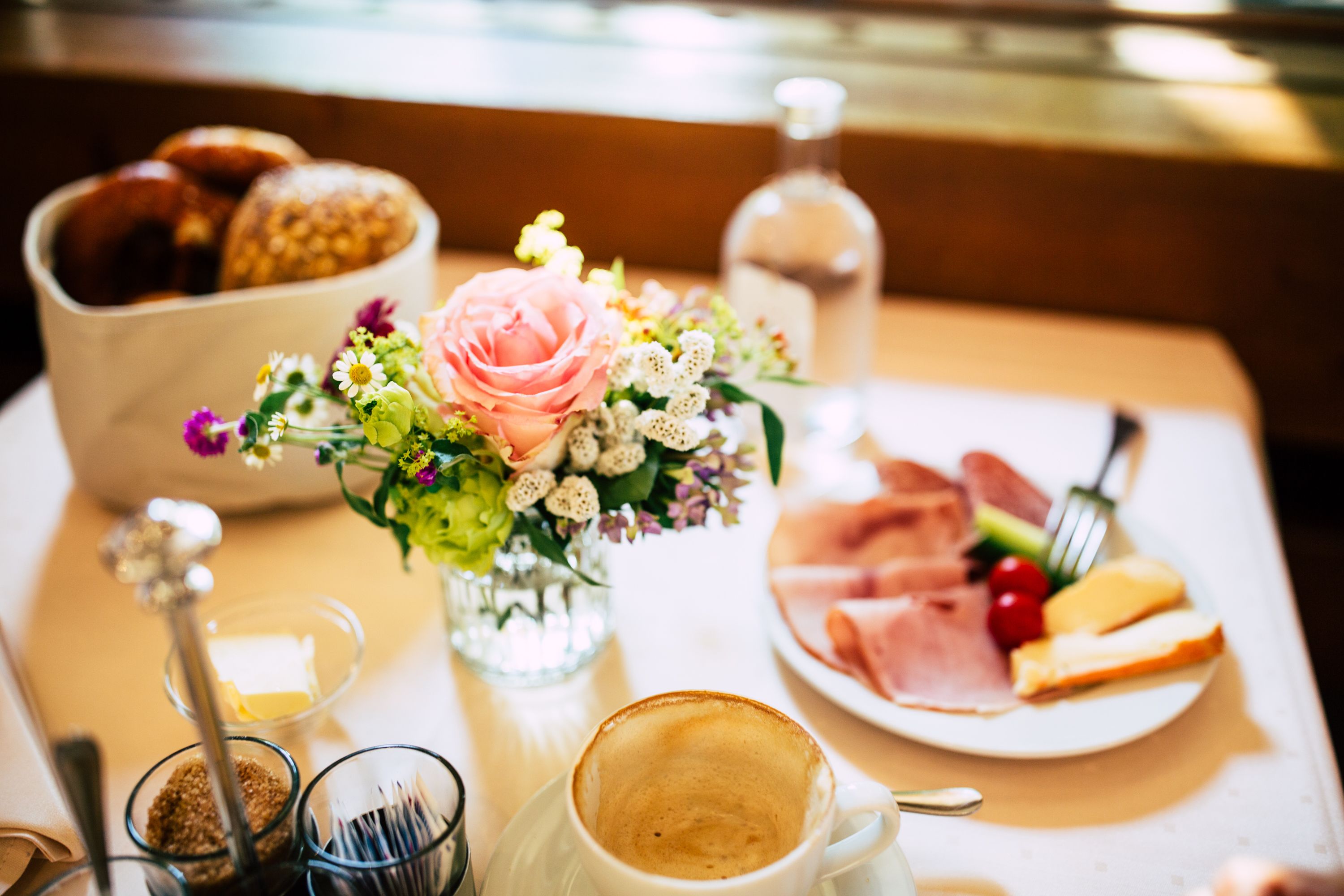 A breakfast setting with a plate of ham, cheese, and cherry tomatoes, a cup of coffee, a glass of milk, a bowl of pastries, and a vase with flowers.
