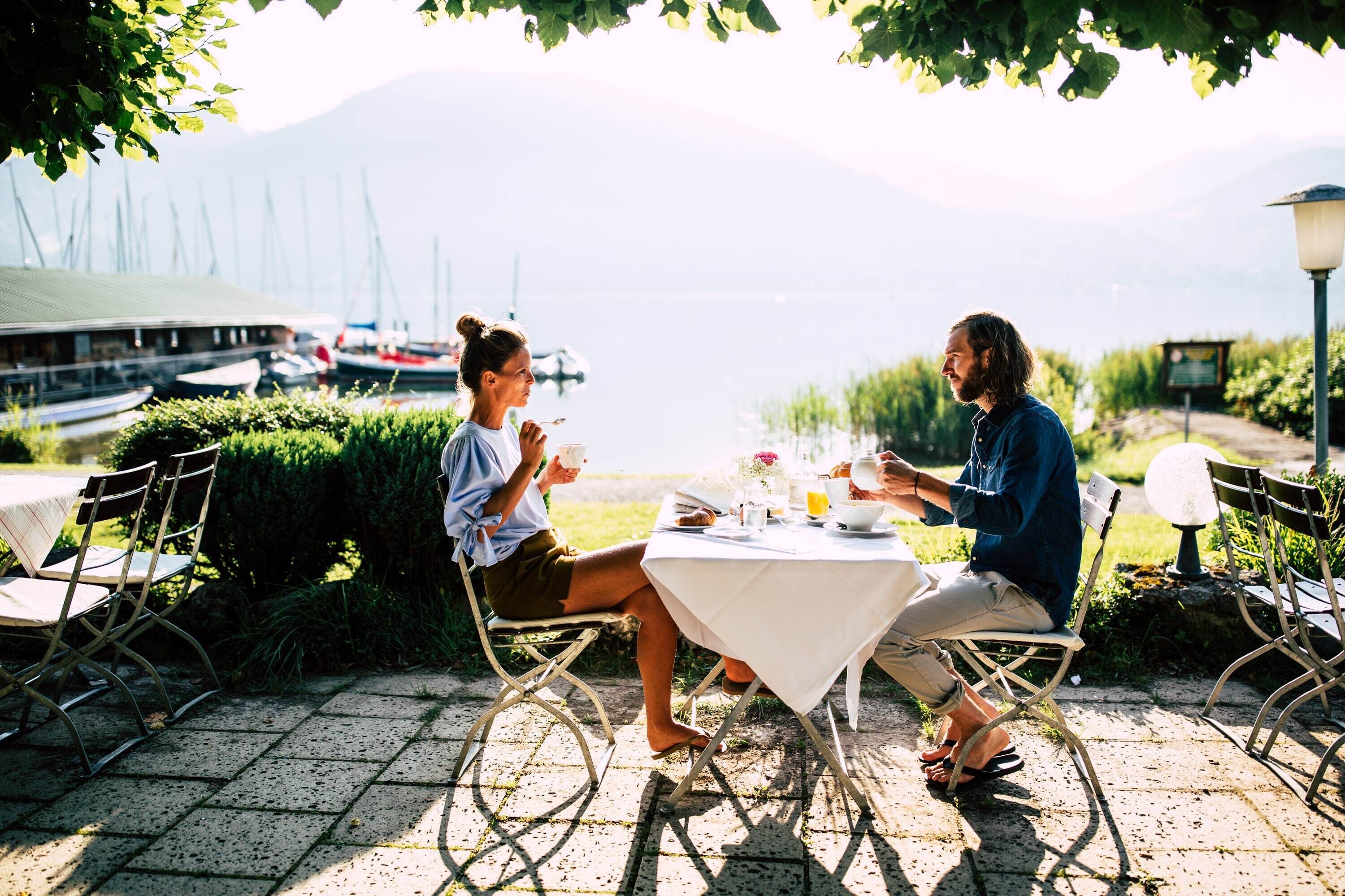 Deux personnes dînent à une table en plein air, avec en toile de fond des bateaux et des montagnes.