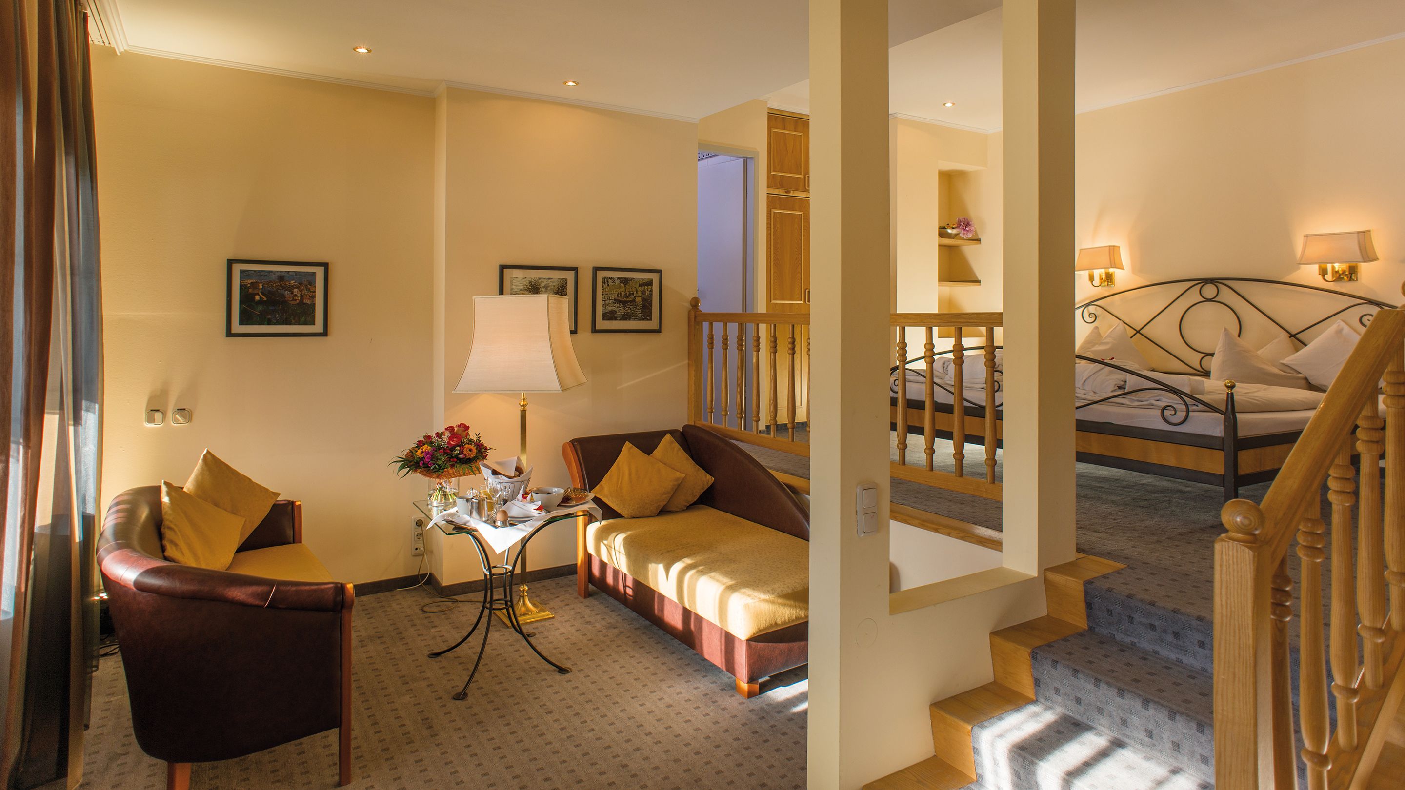 A hotel room with a sofa, armchair, and a small table with a lamp and flowers. There is a staircase leading to another room with a bed.