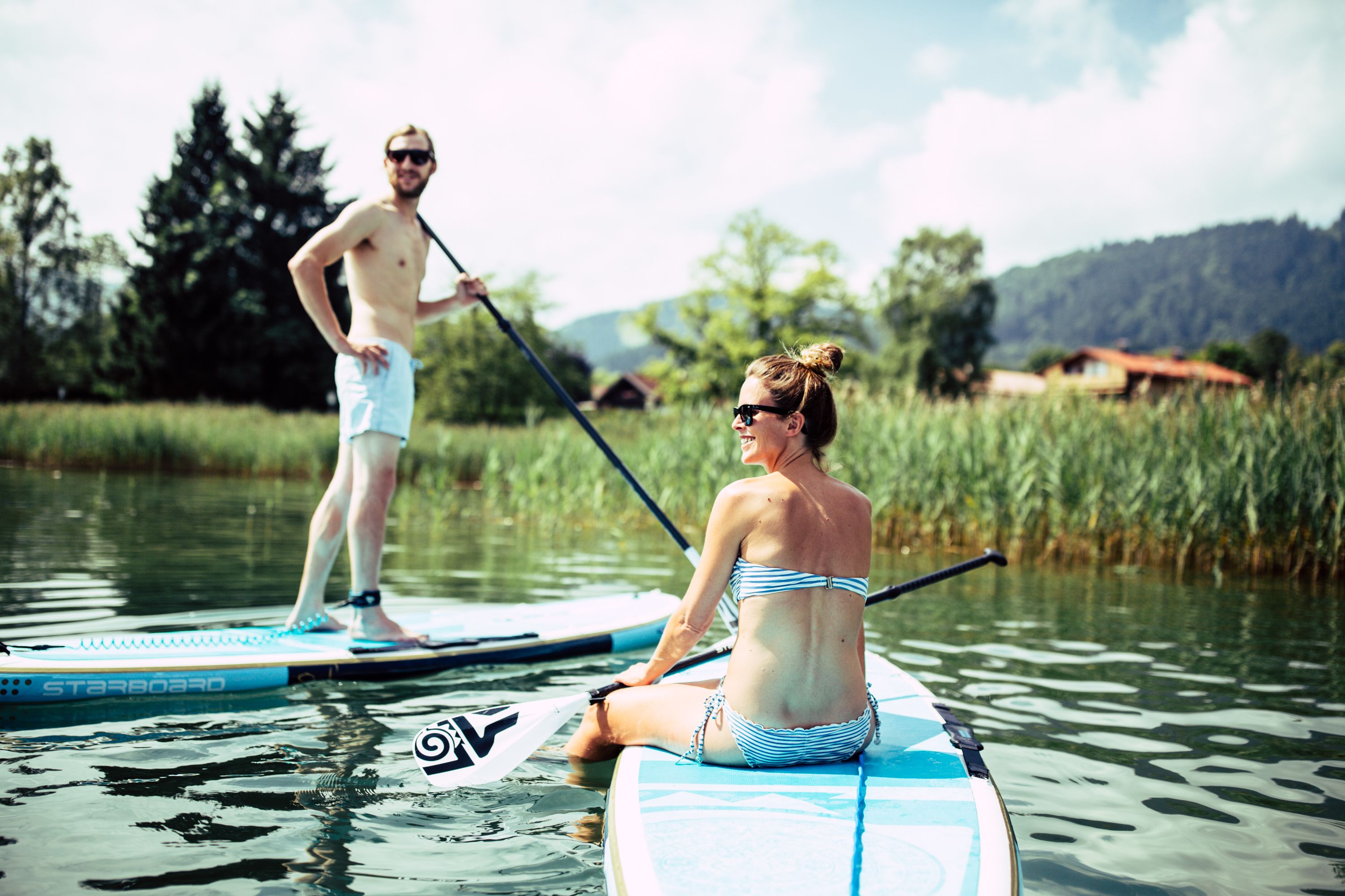 Zwei Personen auf Stand-Up-Boards auf ruhigem Wasser, umgeben von viel Grün und einigen Häusern im Hintergrund.