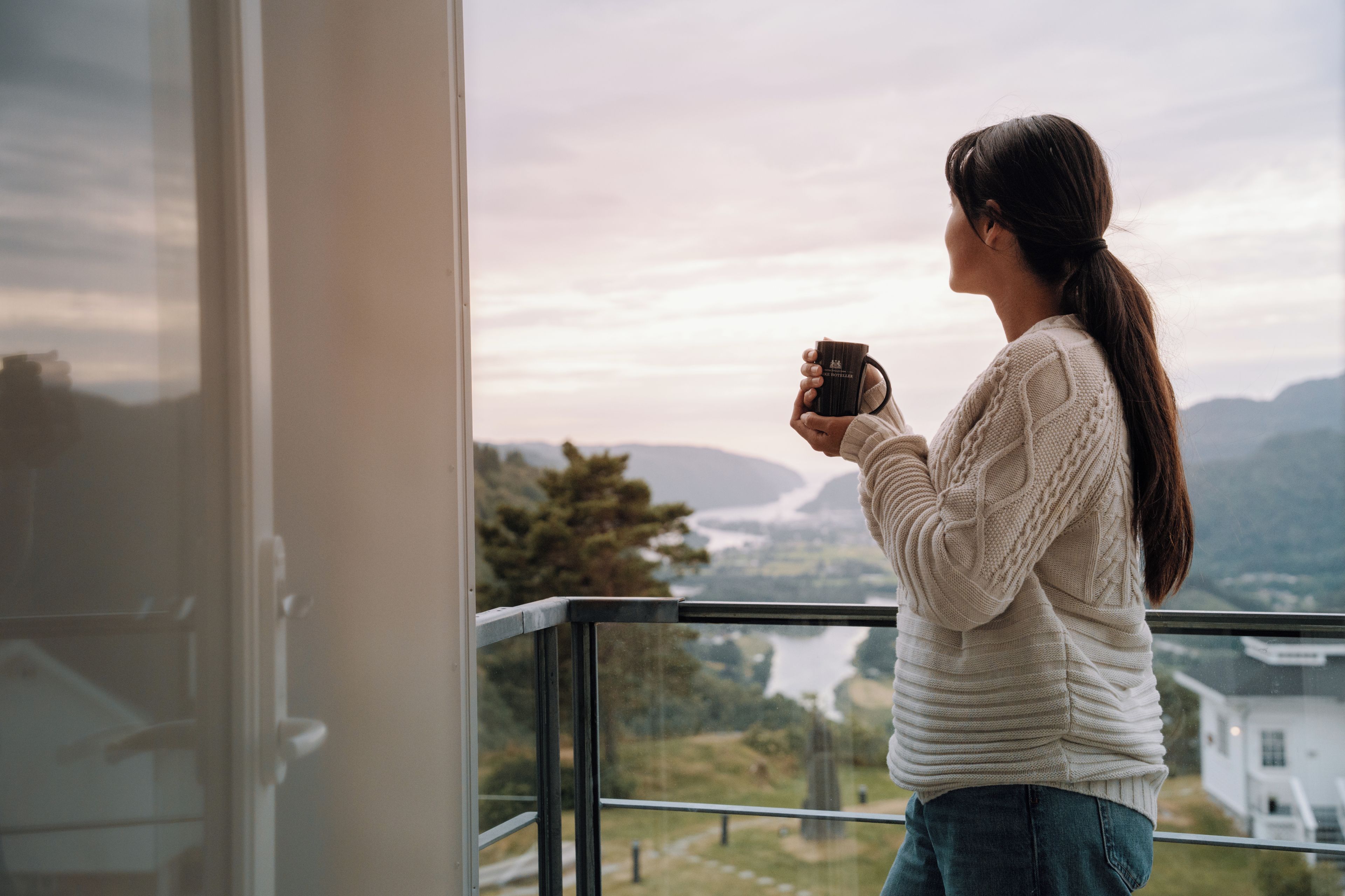 Una persona sosteniendo una taza, de pie junto a una ventana con vistas al paisaje.