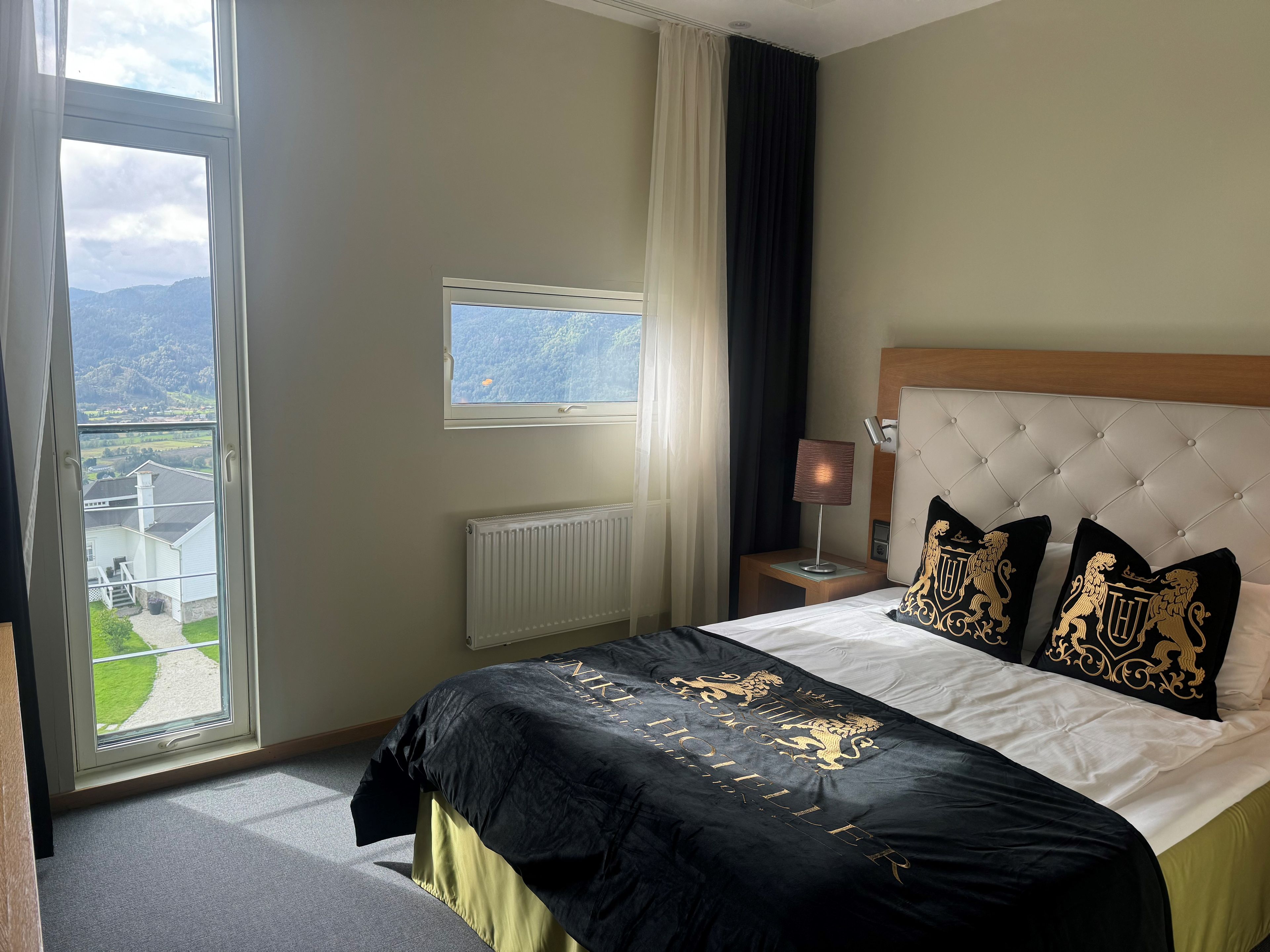 A hotel room with a large bed, a window with a view of the countryside, a bedside table with a lamp, and a radiator.