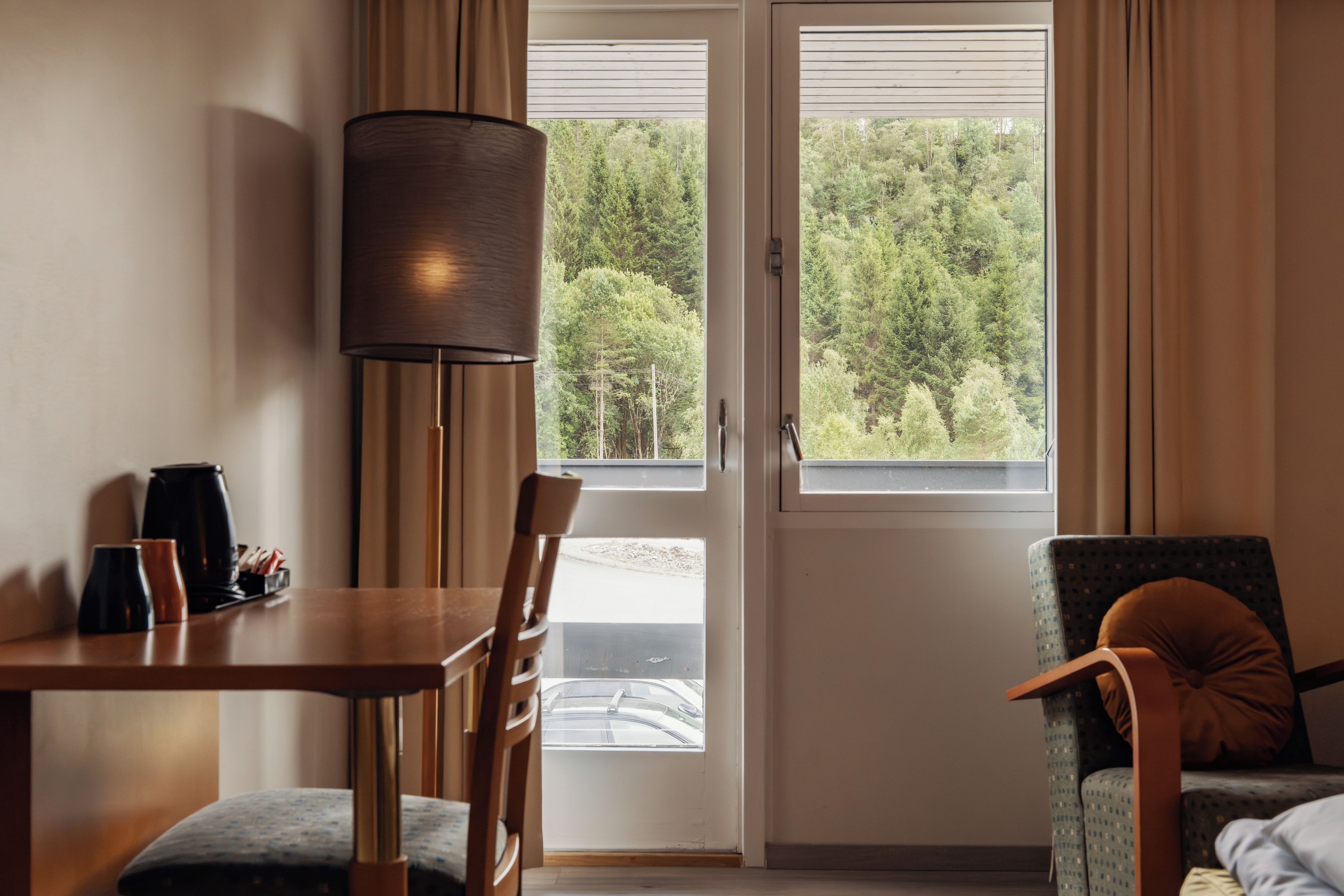 A room with a dining table, chairs, a lamp, a window with a view of trees, and a bed with a pillow.