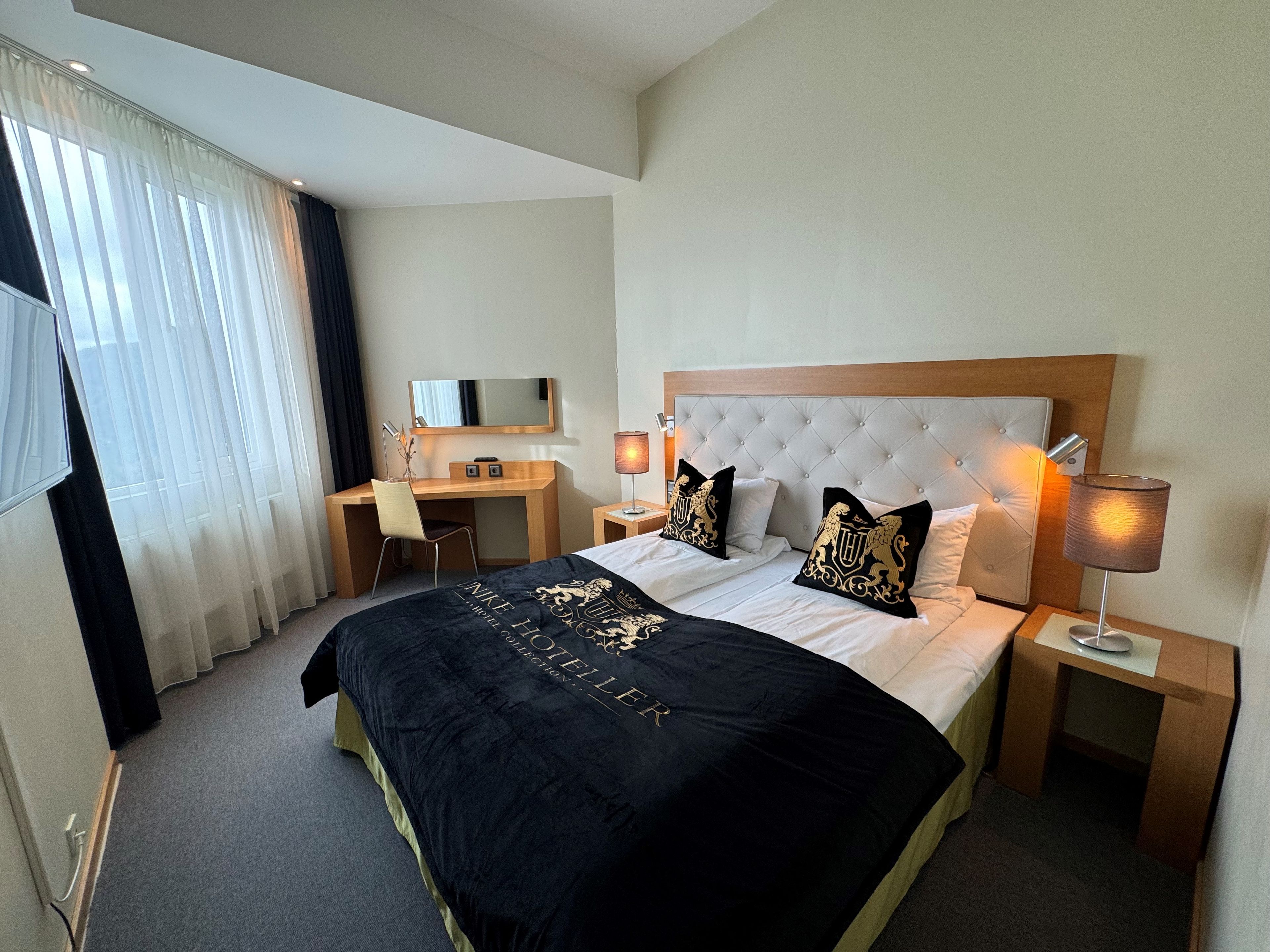 A hotel room with a bed, a desk, a chair, a TV, and a lamp.