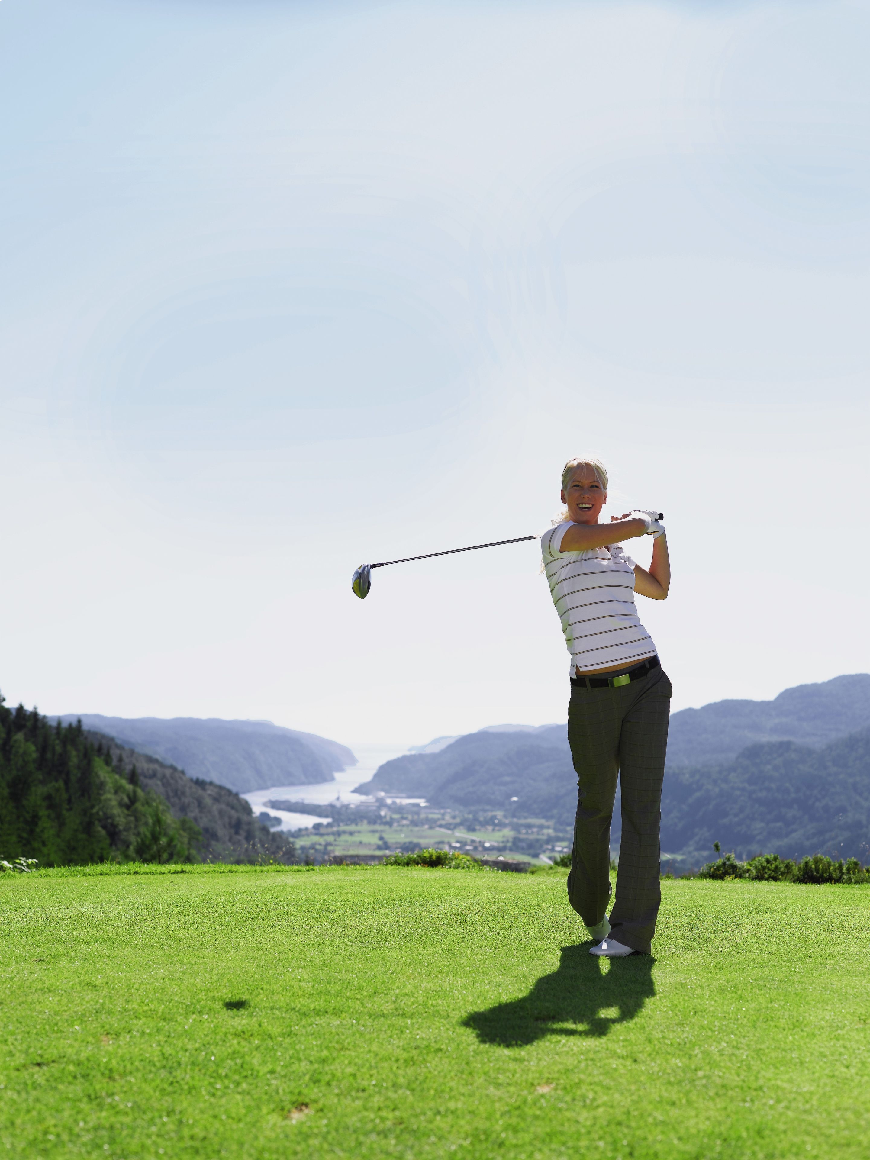 Una persona jugando golf en un campo verde con una vista panorámica de montañas y un río al fondo.