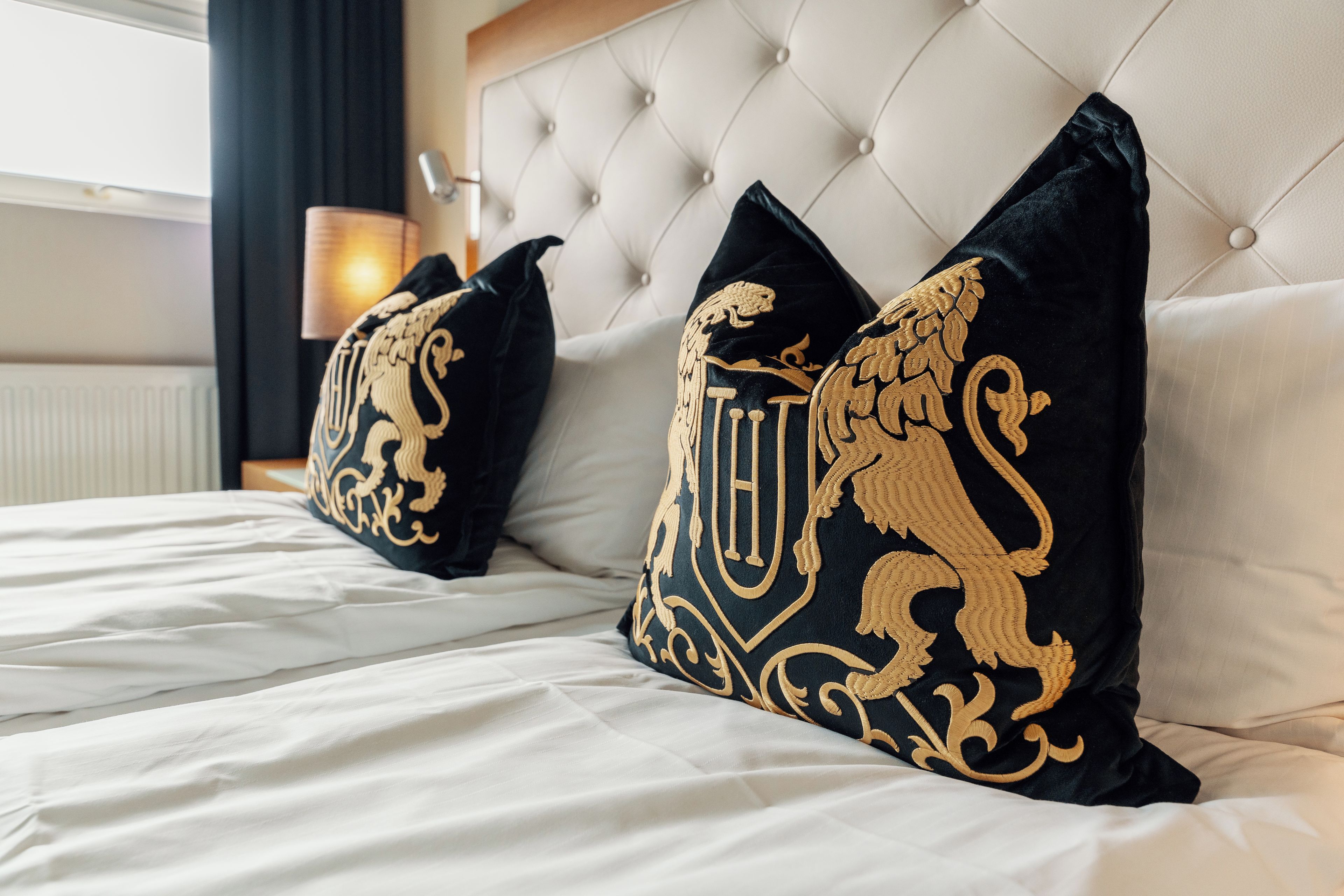 A bed with white bedding and two black pillows with gold embroidery.
