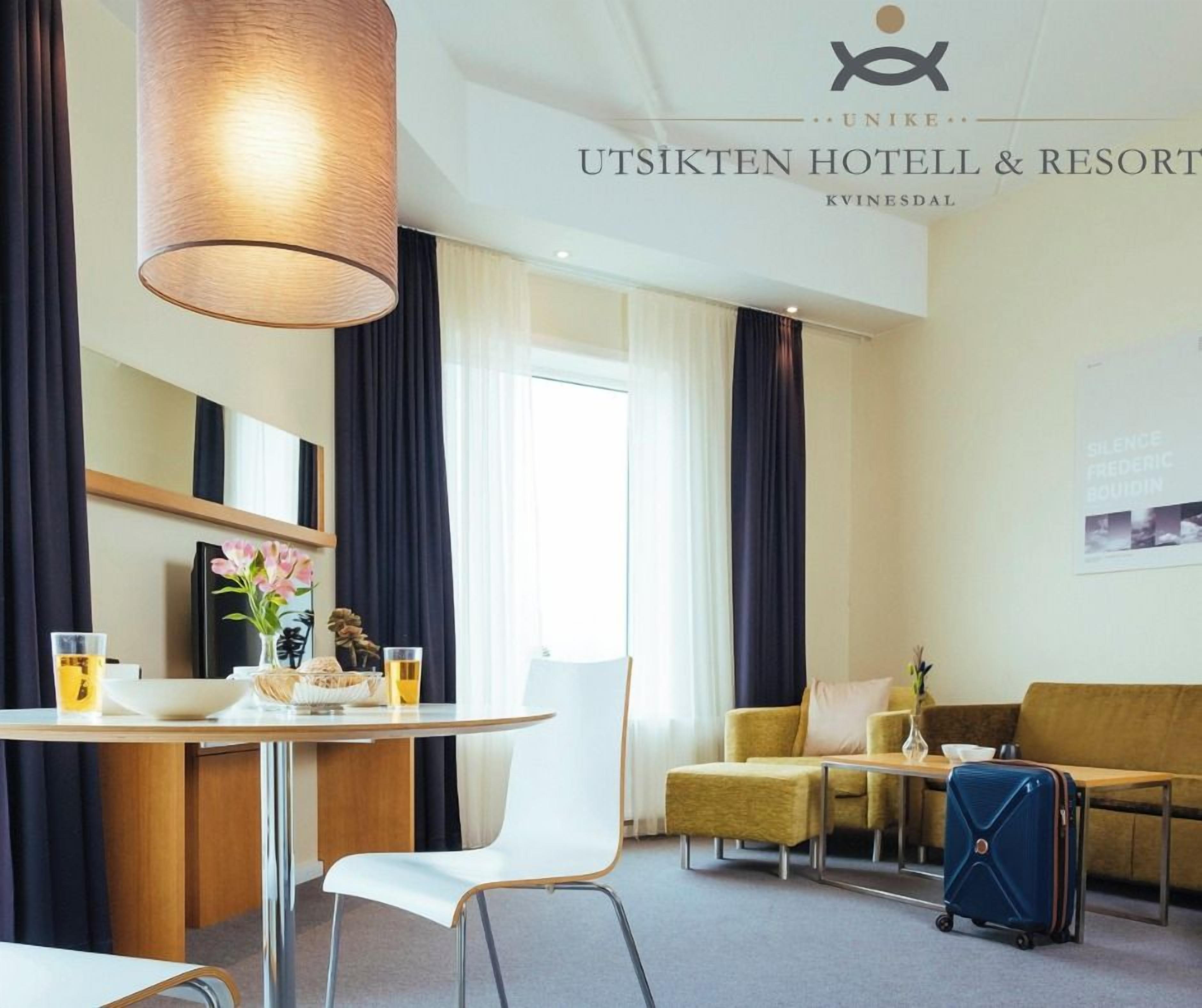 A hotel room with a dining table, a sofa, a chair, a lamp, and a suitcase.