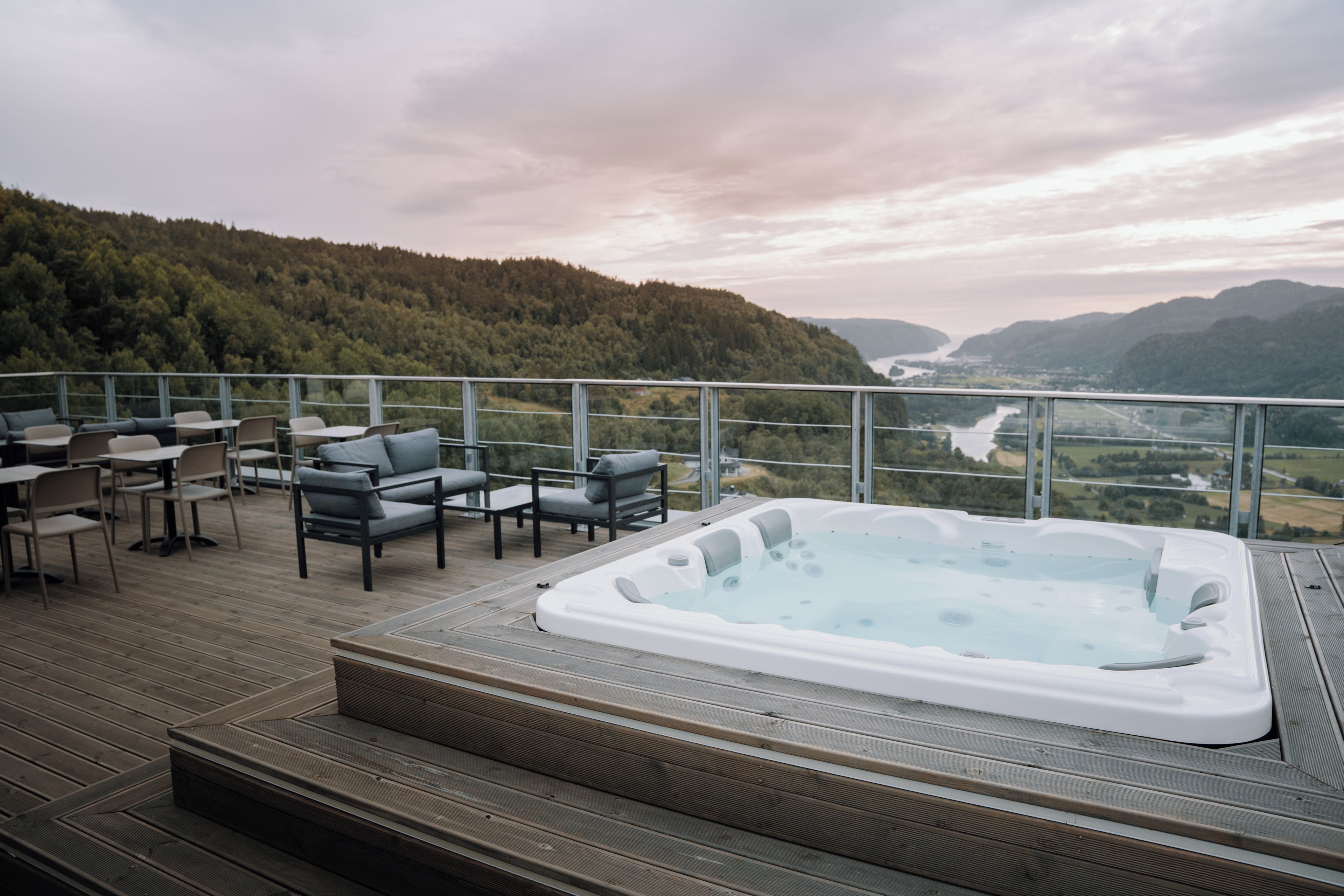 A hot tub on a wooden deck with outdoor seating and a view of a river and mountains.