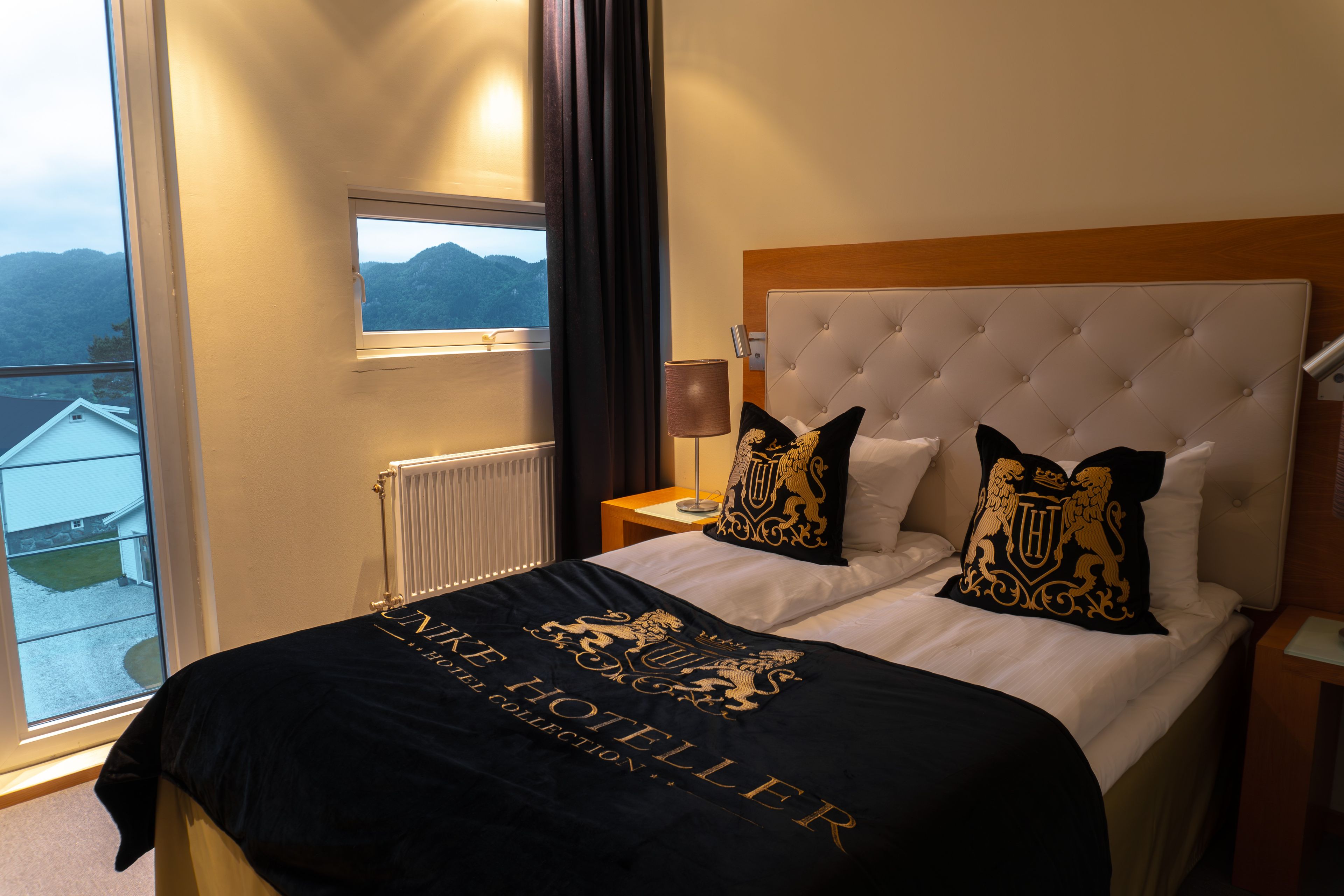 A hotel room with a large window, a bed with a black and gold throw, and a view of the mountains.