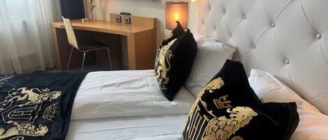 Habitación de hotel con cama, dos almohadas con bordado dorado, un escritorio con silla, una lámpara y un espejo en la pared.