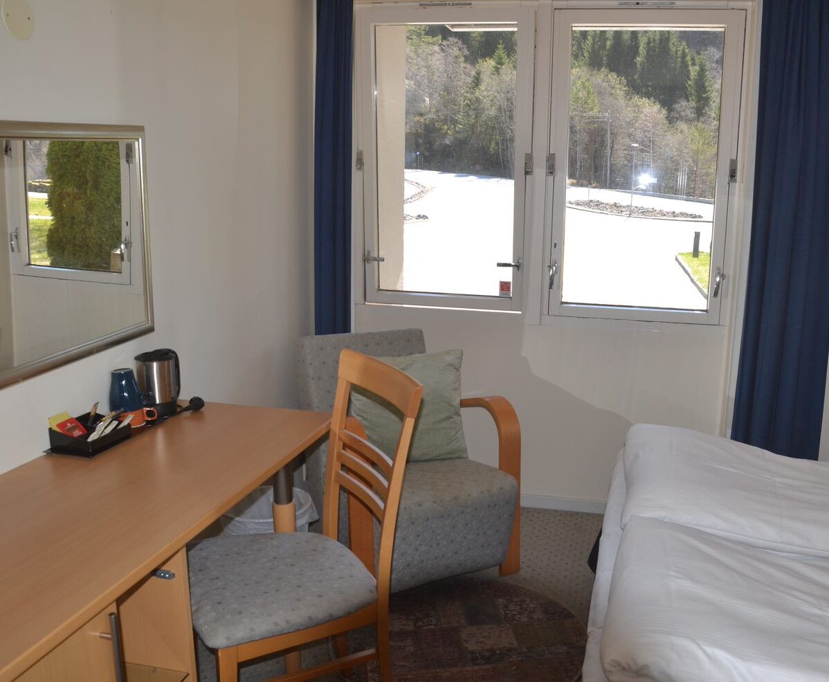 Une chambre d’hôtel avec un lit, un bureau et une chaise, une fenêtre donnant sur un parking et des arbres, et un miroir au mur.