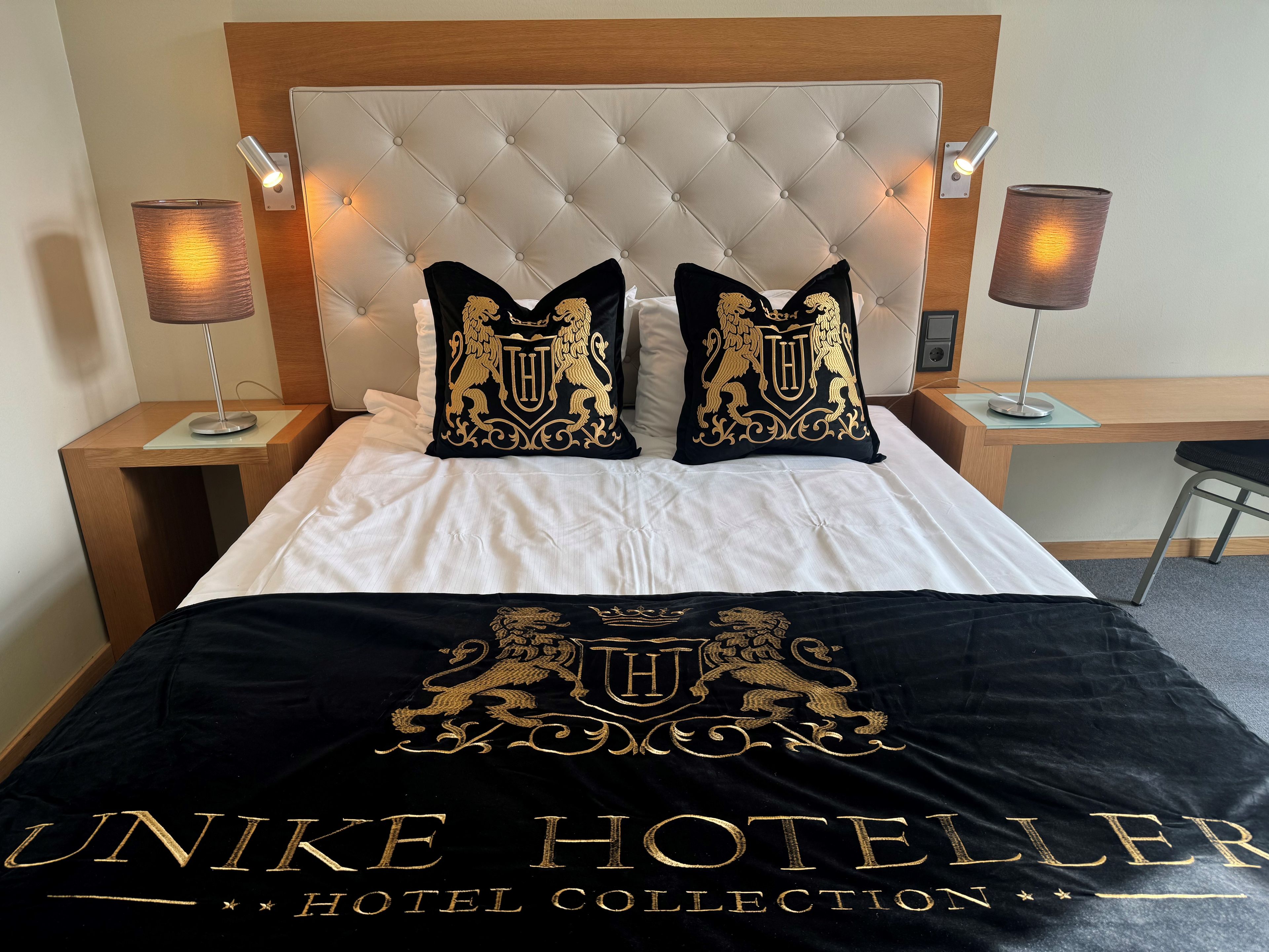 A hotel room with a tufted headboard, two decorative pillows with a crest emblem, a bed with white linens, and a black throw blanket with embossed text.