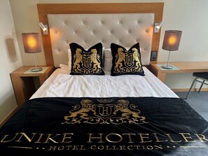 Habitación de hotel con cabecera capitonada, dos almohadas decorativas con un escudo y una cama con ropa blanca, además de un plaid negro con texto en relieve.
