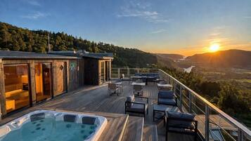 A wooden deck with a hot tub, outdoor seating, and a view of a valley at sunset.