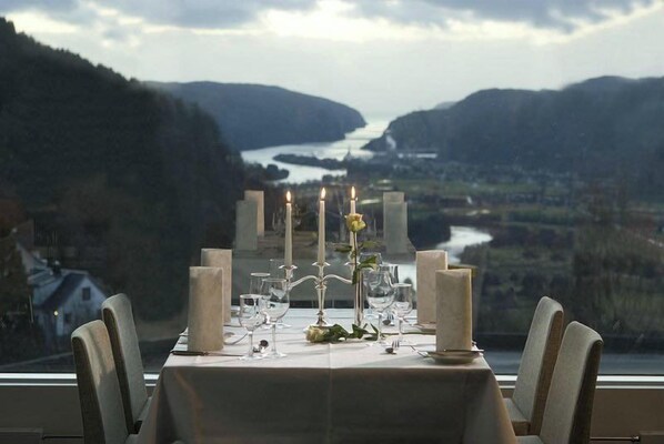 Una mesa preparada para comer, con velas, copas y vistas a un valle y un lago.