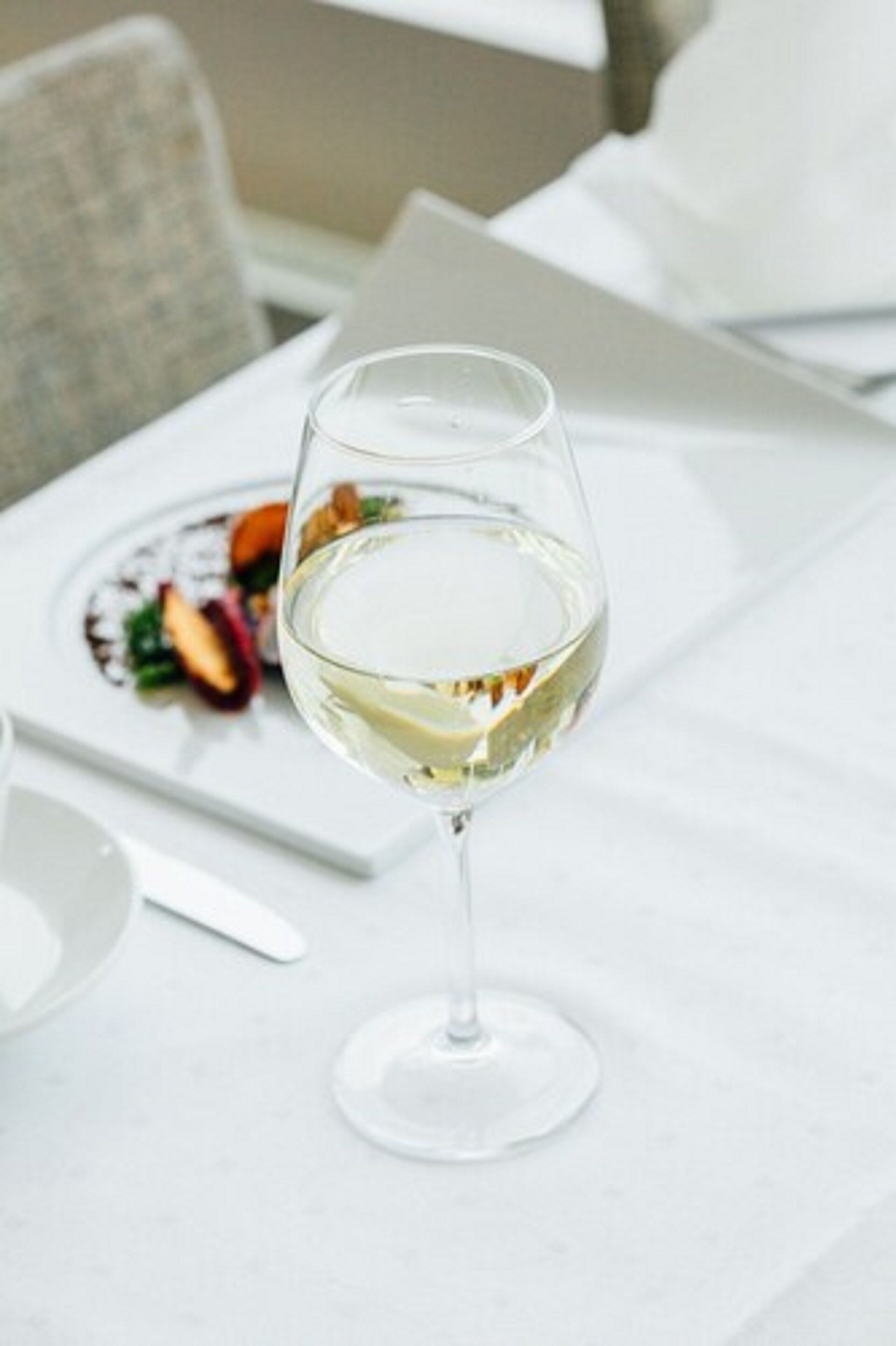Een glas witte wijn op een witte tafel, met een bord eten op de achtergrond.