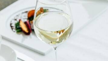 Een glas witte wijn op een witte tafel, met een bord eten op de achtergrond.