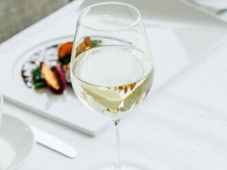 Een glas witte wijn op een witte tafel, met een bord eten op de achtergrond.