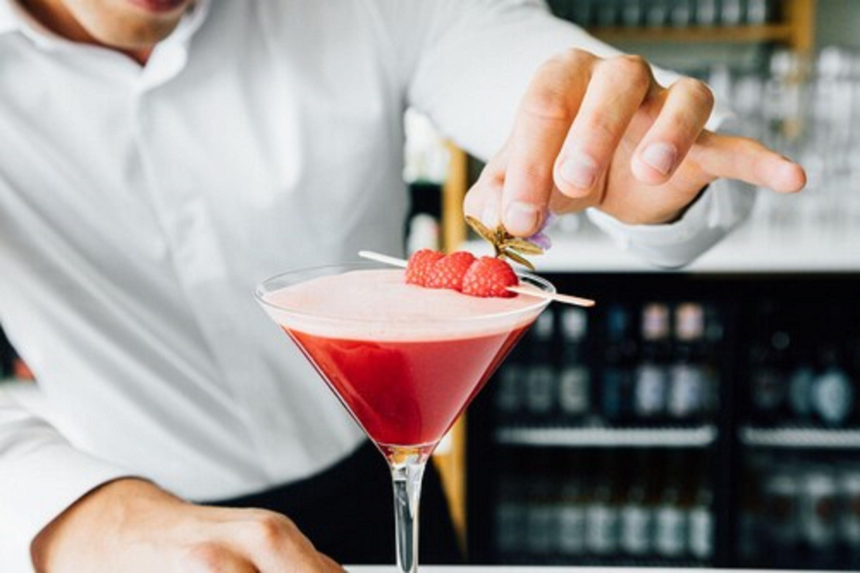 Una persona con camisa blanca señala un cóctel rojo en una copa de martini adornado con una fresa.