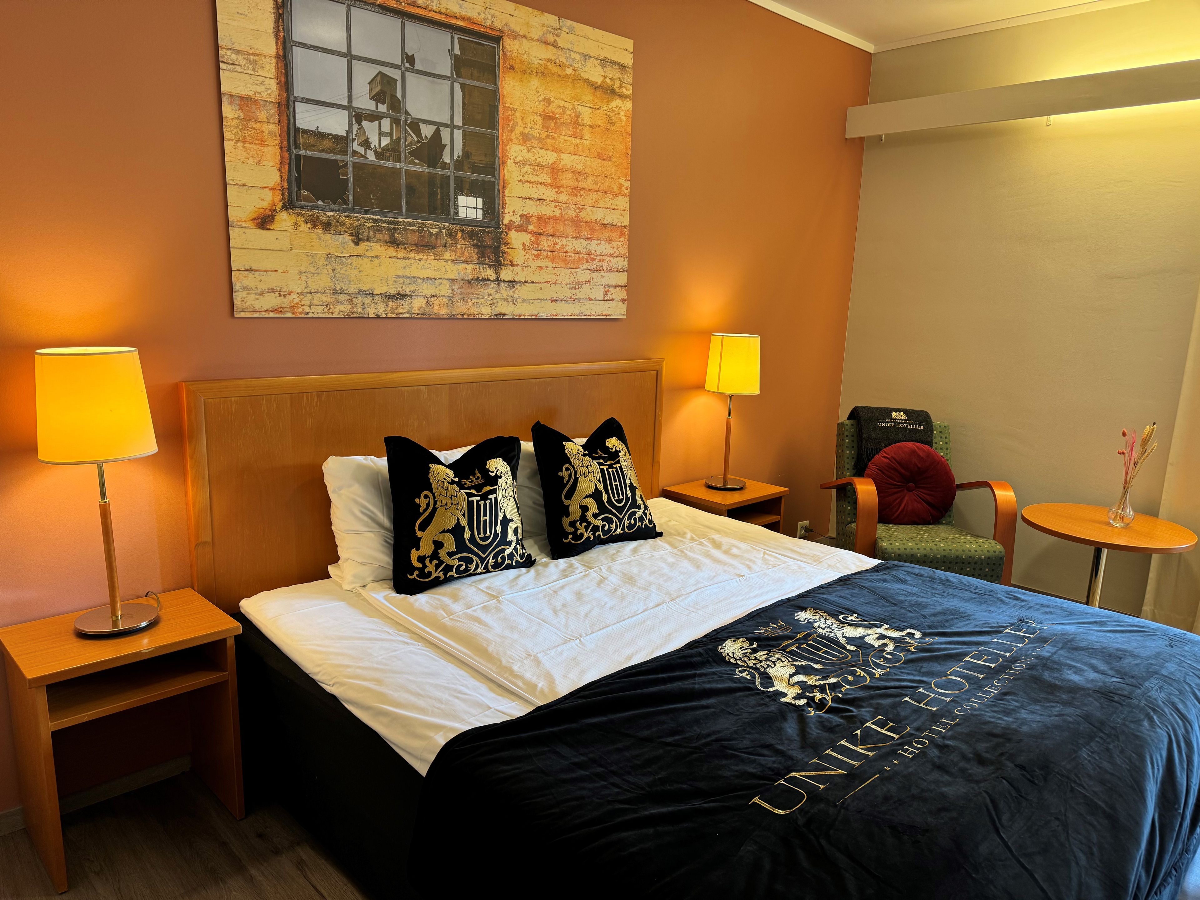 A hotel room with a bed, two bedside lamps, a chair, a small table, and a wall-mounted artwork.