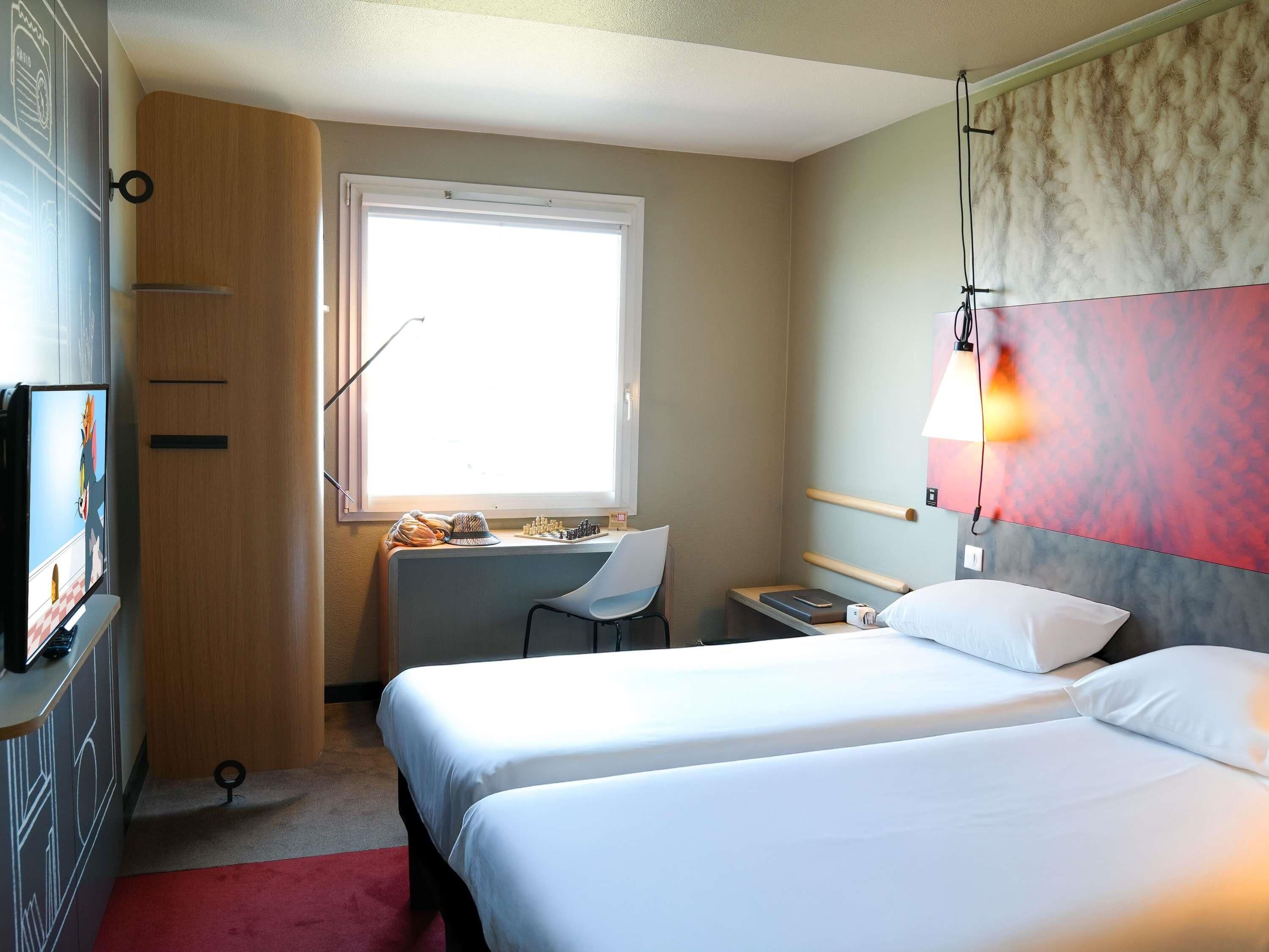 A hotel room with two beds, a desk, a chair, a TV, and a window.