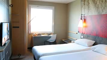 A hotel room with two beds, a desk, a chair, a TV, and a window.