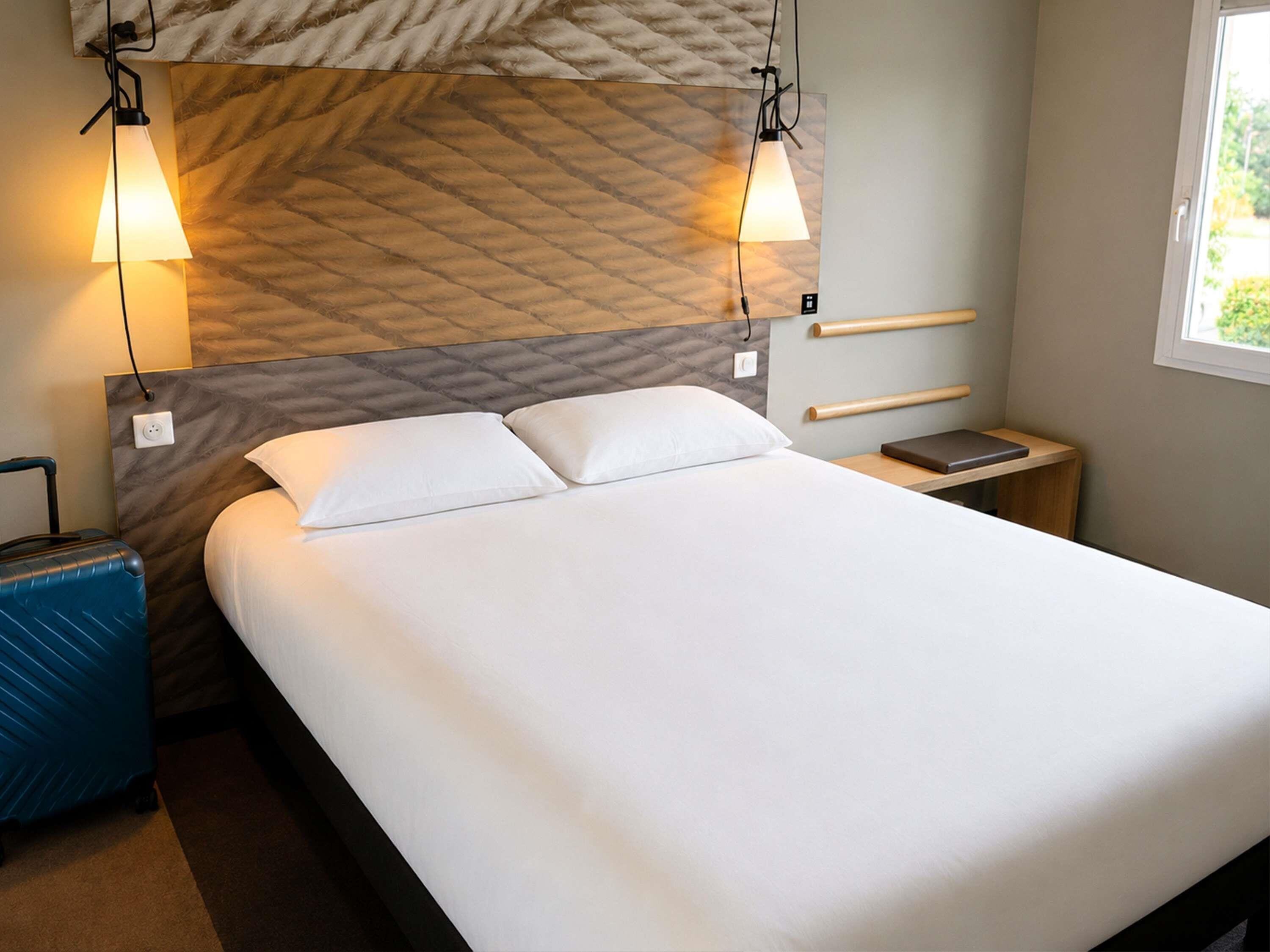 Une chambre d’hôtel avec un lit, des tables de chevet, une valise et une fenêtre.