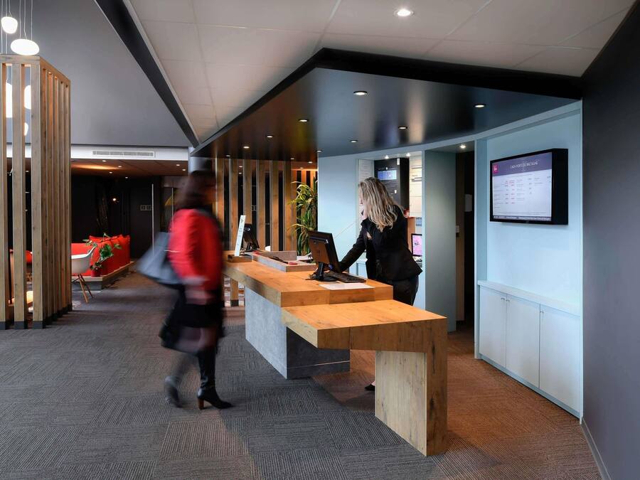 A modern reception desk with a computer, a seating area with red cushions, and a wall-mounted information board.