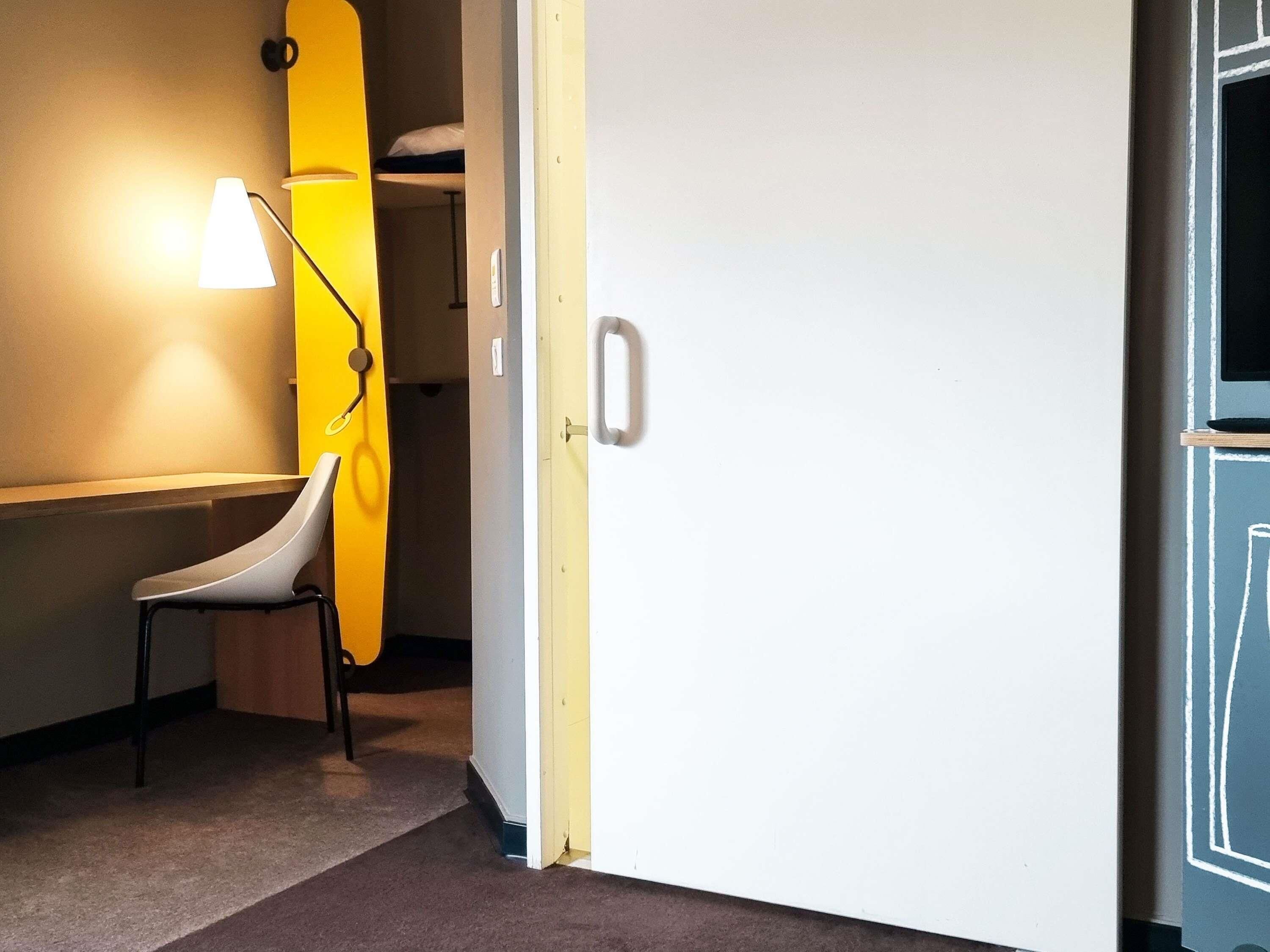 Una habitación con una puerta blanca, una silla blanca, una tabla de surf amarilla apoyada en la pared y una lámpara.