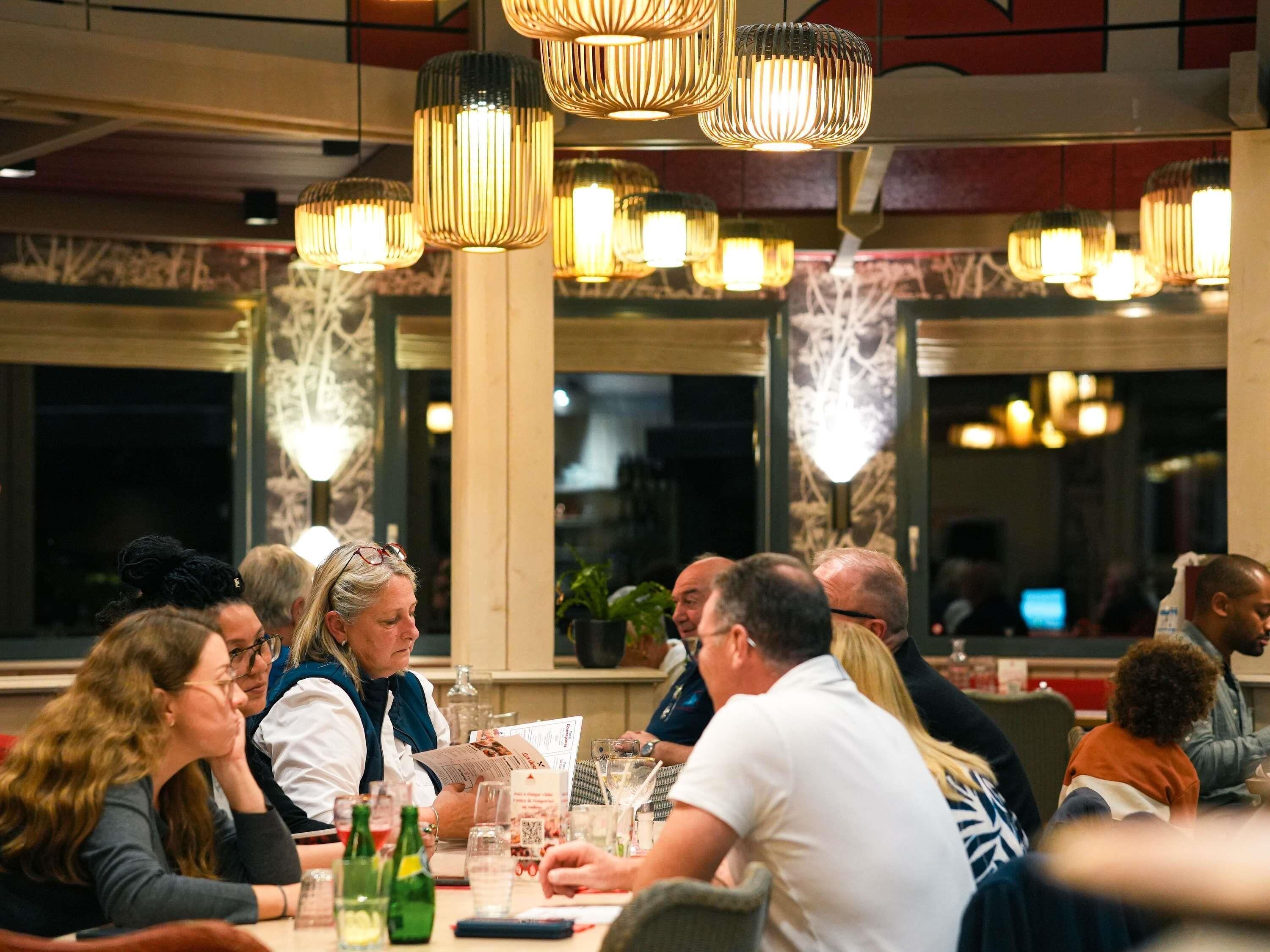 Un groupe de personnes est réuni autour d’une table dans un restaurant. Des lampes suspendues illuminent la salle, et de grandes fenêtres offrent une vue sur le paysage urbain.