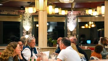 Un groupe de personnes est réuni autour d’une table dans un restaurant. Des lampes suspendues illuminent la salle, et de grandes fenêtres offrent une vue sur le paysage urbain.
