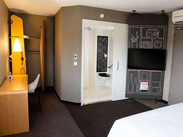 A hotel room with a bed, a desk with a lamp, a shower, a toilet, and a TV mounted on the wall.