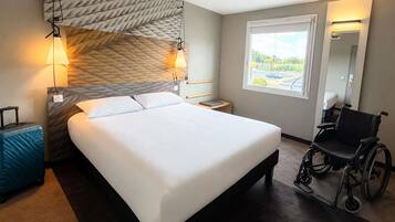 A hotel room with a large bed, a wheelchair, a suitcase, and a window with a view of the outside.