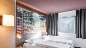 A hotel room with a bed, a nightstand, a lamp, a window with curtains, and a wall with departure information. - B&B HOTEL Hamburg-Airport (Hamburg)