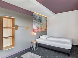 Ein modernes Hotelzimmer mit einer grauen Couch, einem Holzschrank und einem an der Wand montierten Fernseher, der Abfluginformationen anzeigt.