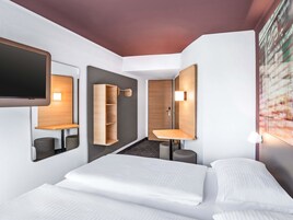 Ein modernes Hotelzimmer mit einem Bett, einem integrierten Fernseher, einem Holzschrank, einem Spiegel und einem an der Wand befestigten Regal.
