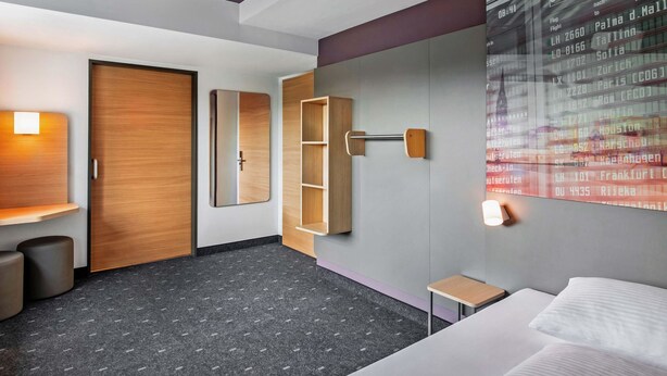 Una habitación de hotel moderna con cama, escritorio, armario y un televisor de pared que muestra información de vuelos.