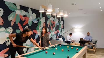 A pool table with balls, a person playing, and another person observing. There are ceiling lights and a colorful wall mural.